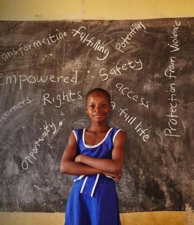 A young girl stands in her school with words written on a blackboard