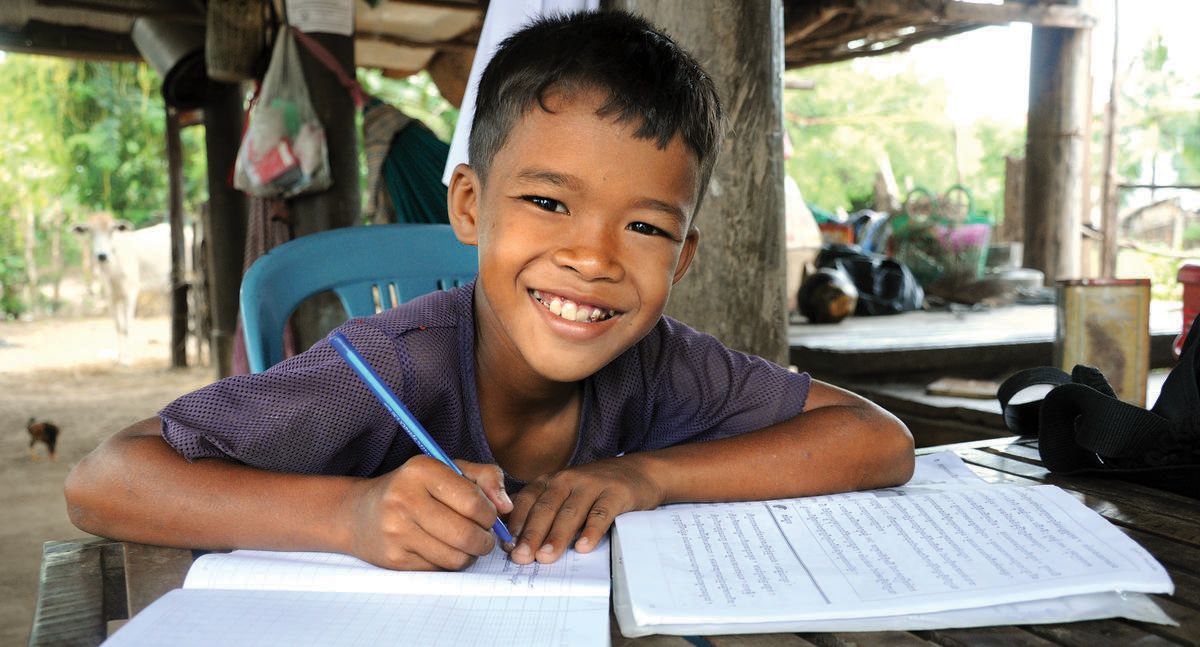 Khmer boy smiles at the camera with pen in hand, writing in his school books, Cambodia.