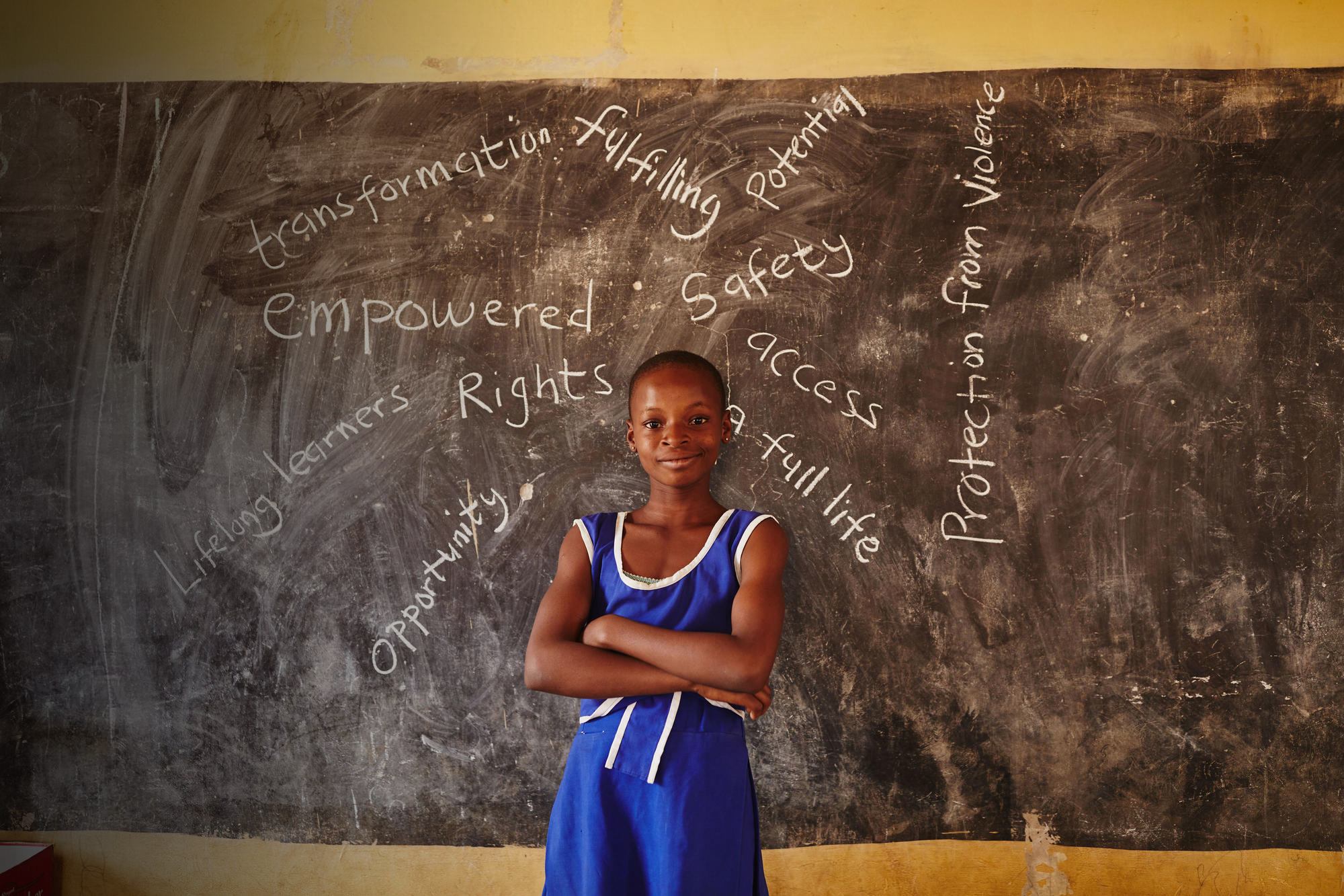 A young girl stands in her school with words written on a blackboard
