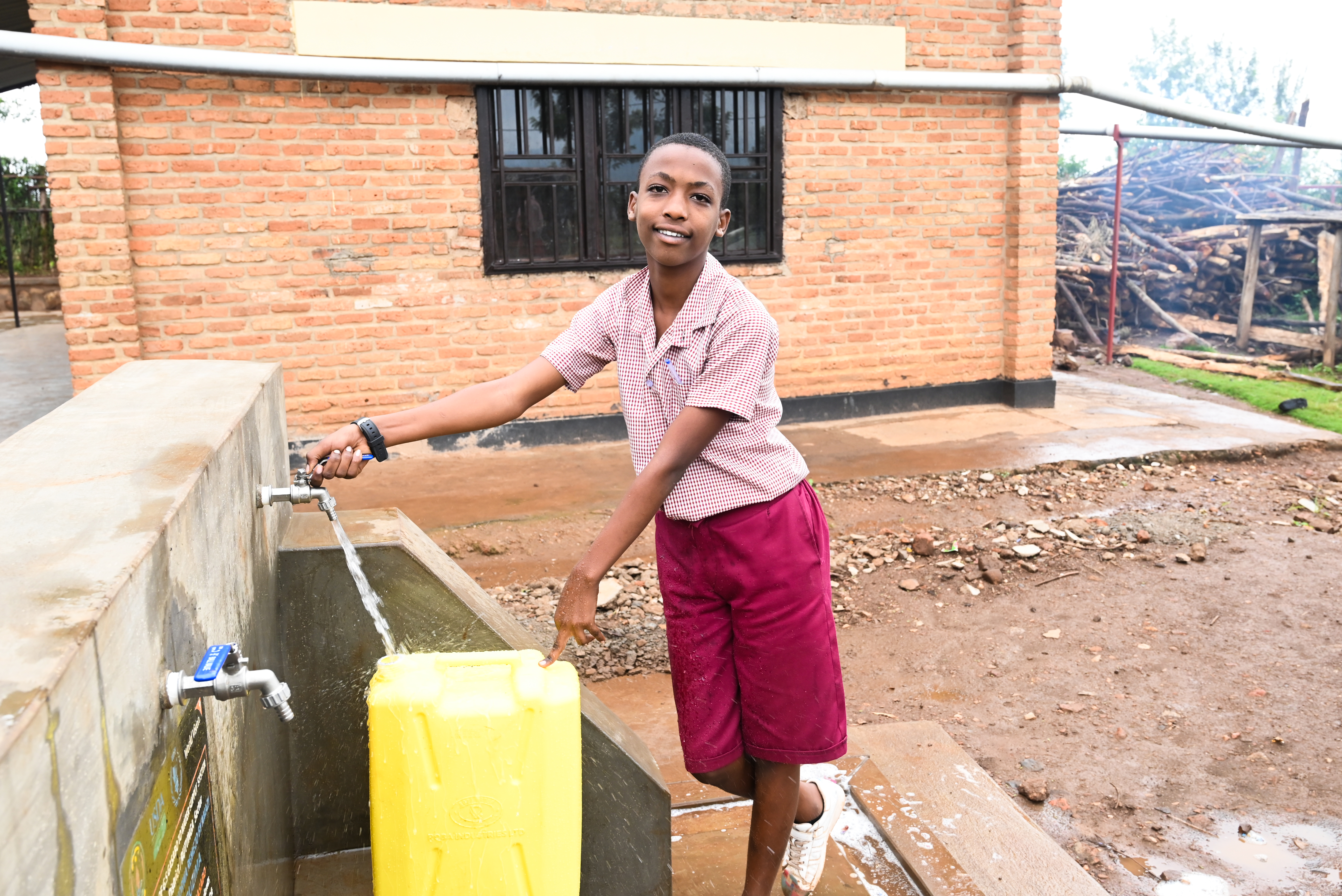 A Young boy in a school uniform collects water in a yellow container at a school water point. 