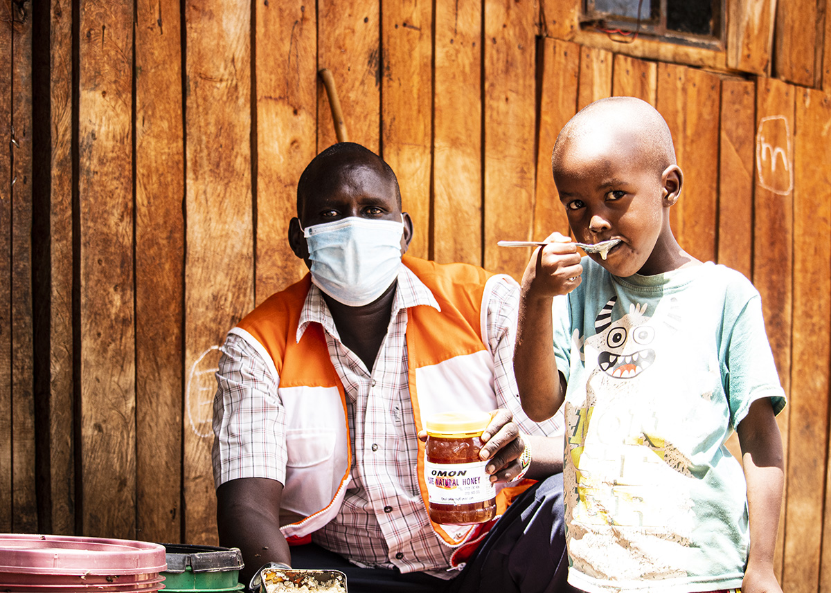 Titus Ng’otiek eats honey from a spoon as World vision’s Philip Ole Molo looks on.