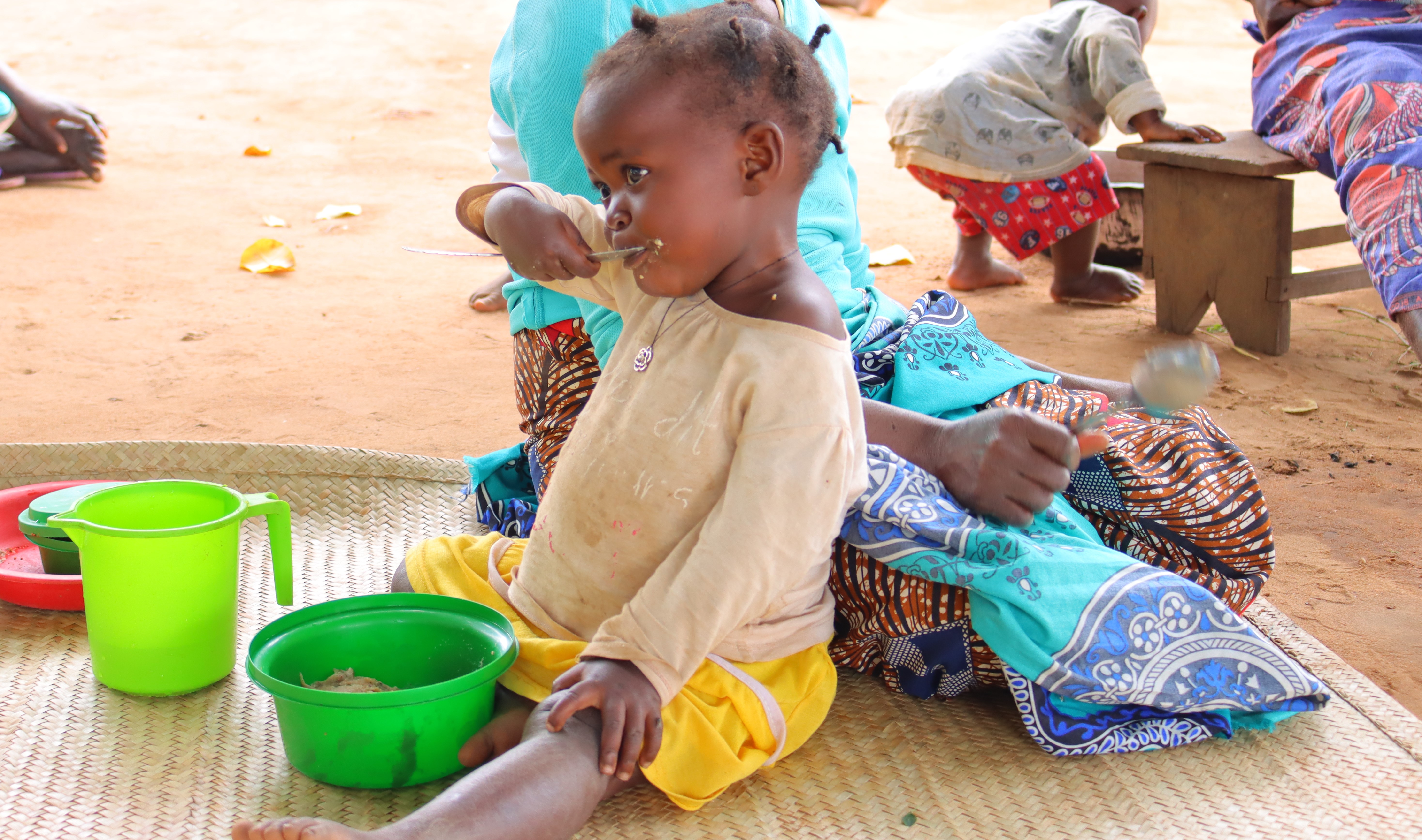 Anatércia, seated in a straw mat, alongside her mother, eats and enjoys her fortified porridge 