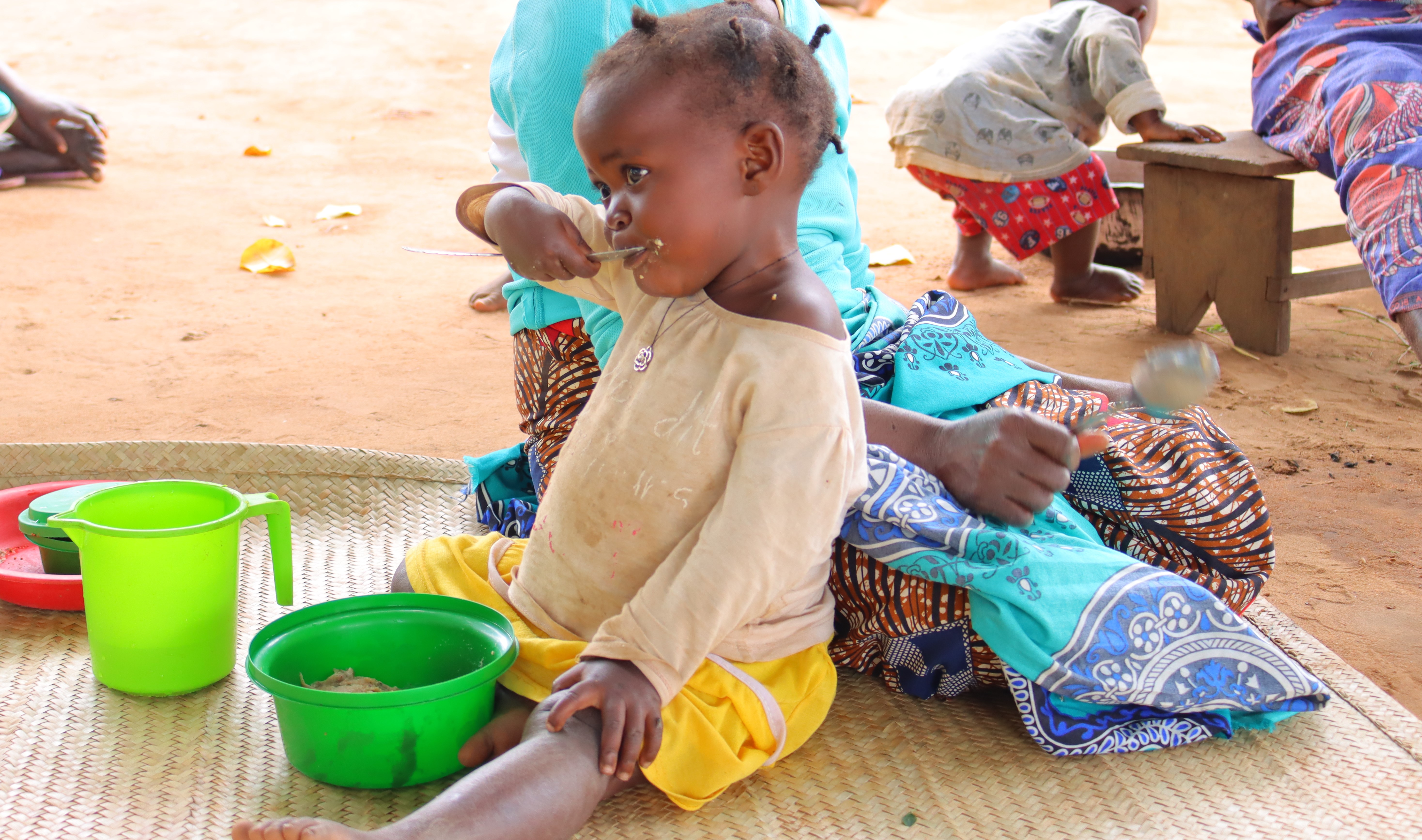 Anatércia, seated in a straw mat, alongside her mother, eats and enjoys her fortified porridge