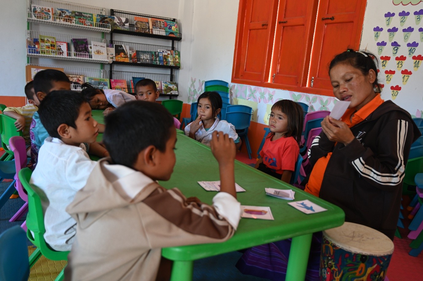 A teacher sits at a low green table with a group of young children in a classroom.