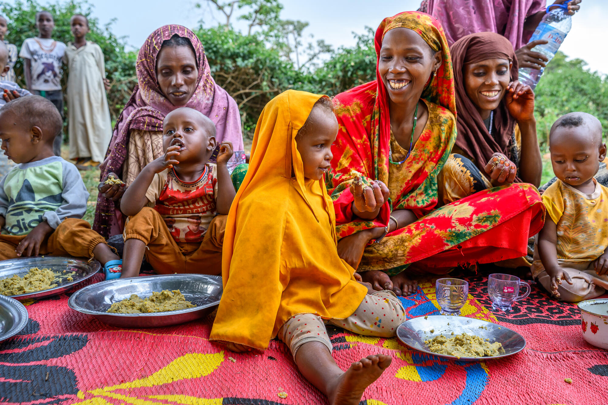 Three women sit on a mat feeding small children porridge from silver colored plates.