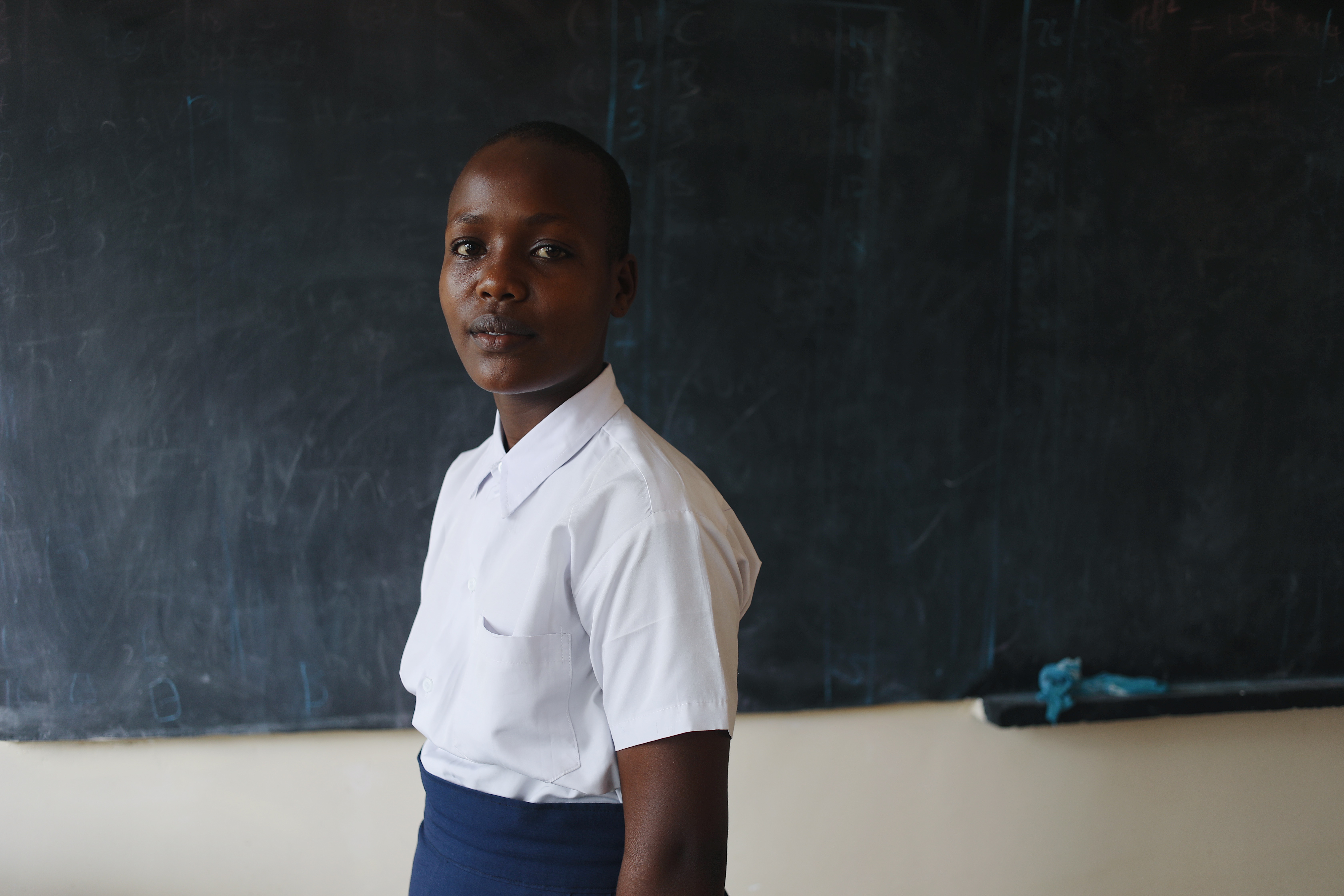 Ester, 16, poses for a photo in front of the blackboard at her school