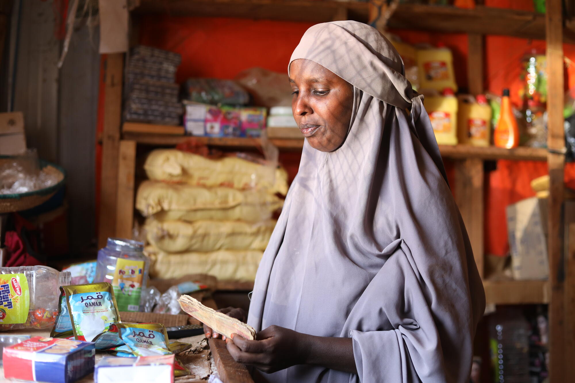 A woman wearing a grey hijab stands inside her shop looking down at money she's holding in her hand.