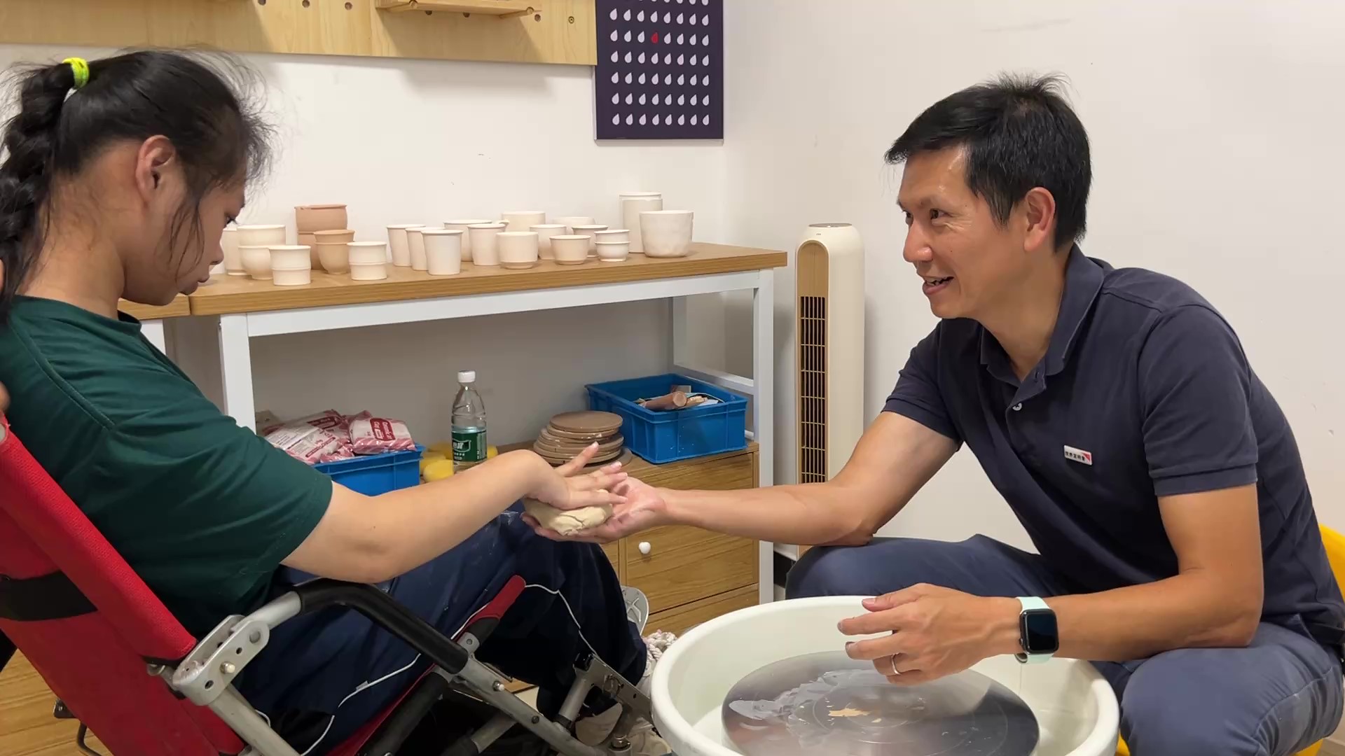 A man sits with a young girl in a wheel chair in a pottery studio.