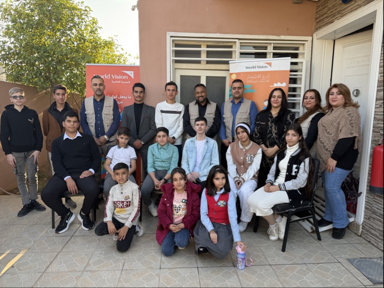 A group of children and World Vision staff sit and stand for a photo in front of 2 World Vision banners