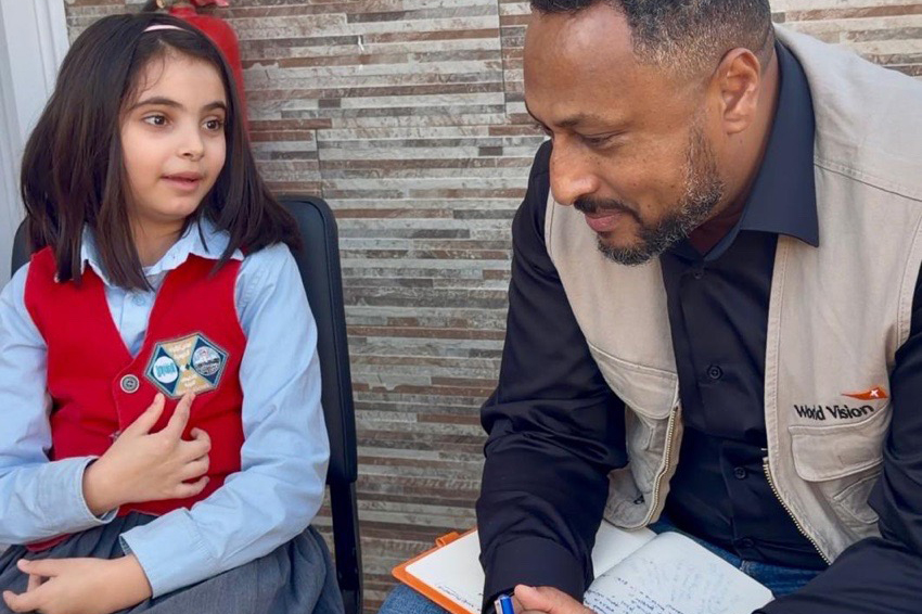 A young girls sits in a chair next to a Work Vision male staff who listens to her talk.