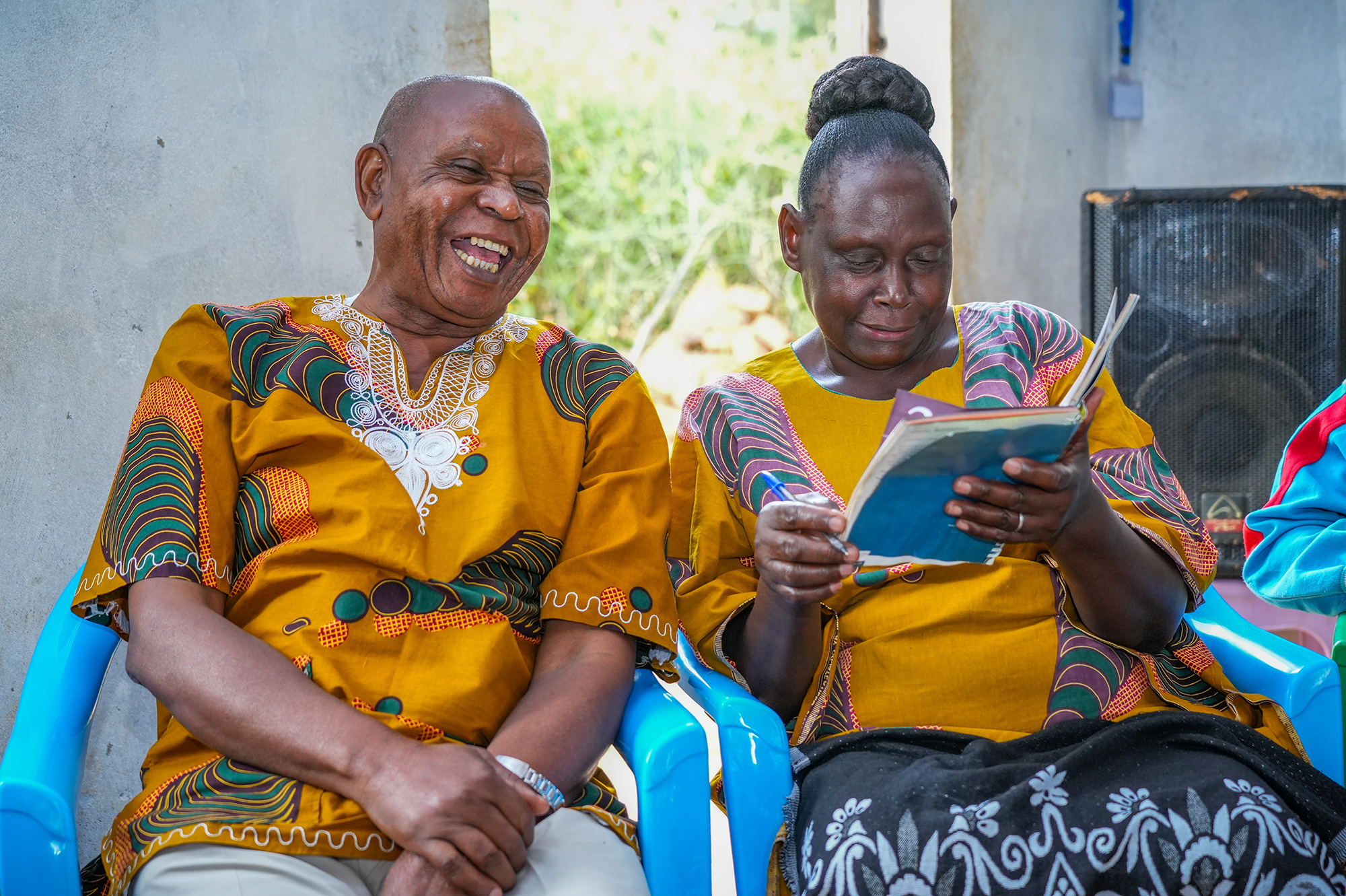 A man and woman sit in blue plastic chairs. The man is smiling big while the woman looks down at a book in her hands.
