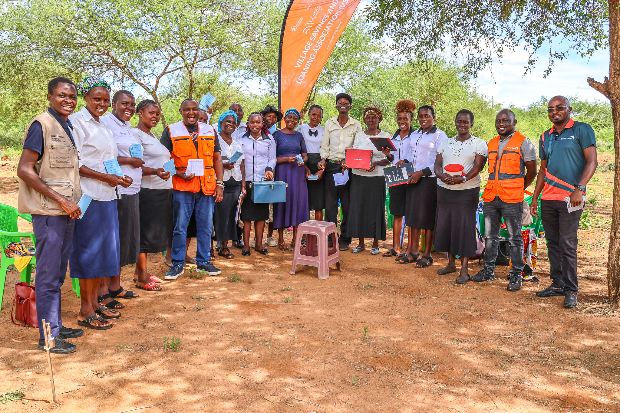 World Vision Kenya staff with Mwanzo Mpya VSLA members after a weekly table banking session in Kyase Village, Makueni County. Members proudly hold their savings box and record books. © World Vision Kenya/ Jared Ontobo