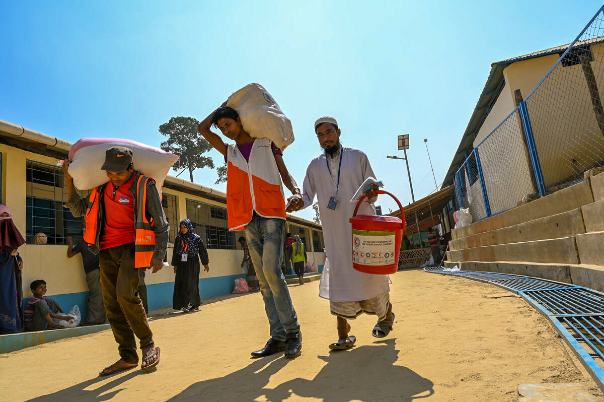 A fellow Rohingya porter and a volunteer helps Khaled to carry his food from the e-voucher shop back to his shelter.