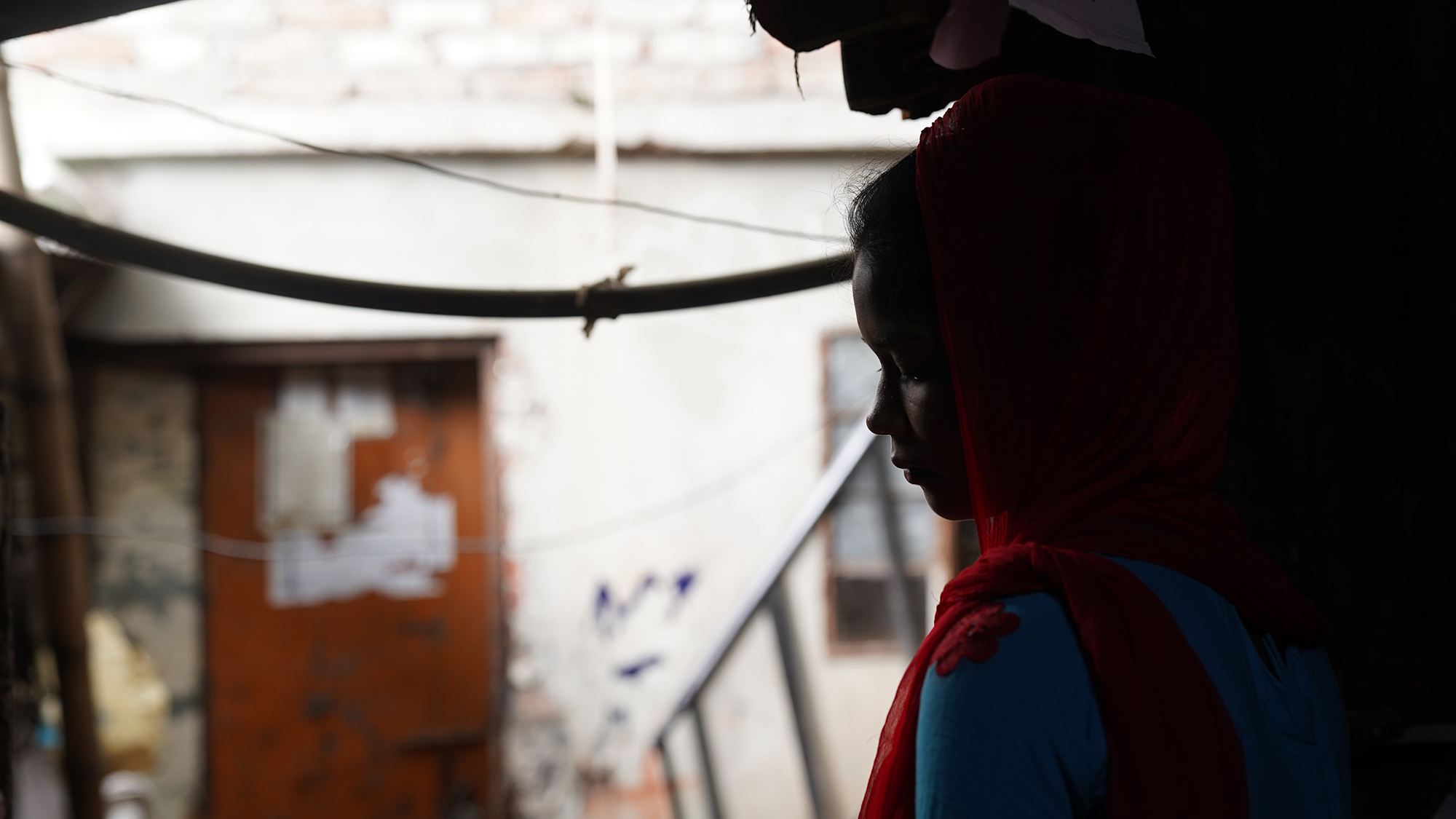 a young girl with a red head scarf stares sadly out a window in a slum settlement.