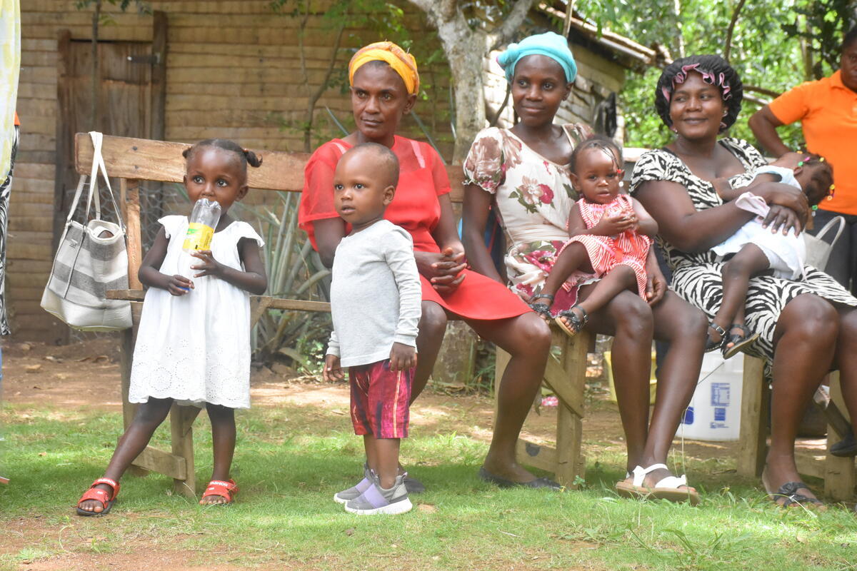 Mothers with small children sit on bench in grassy field in Haiti