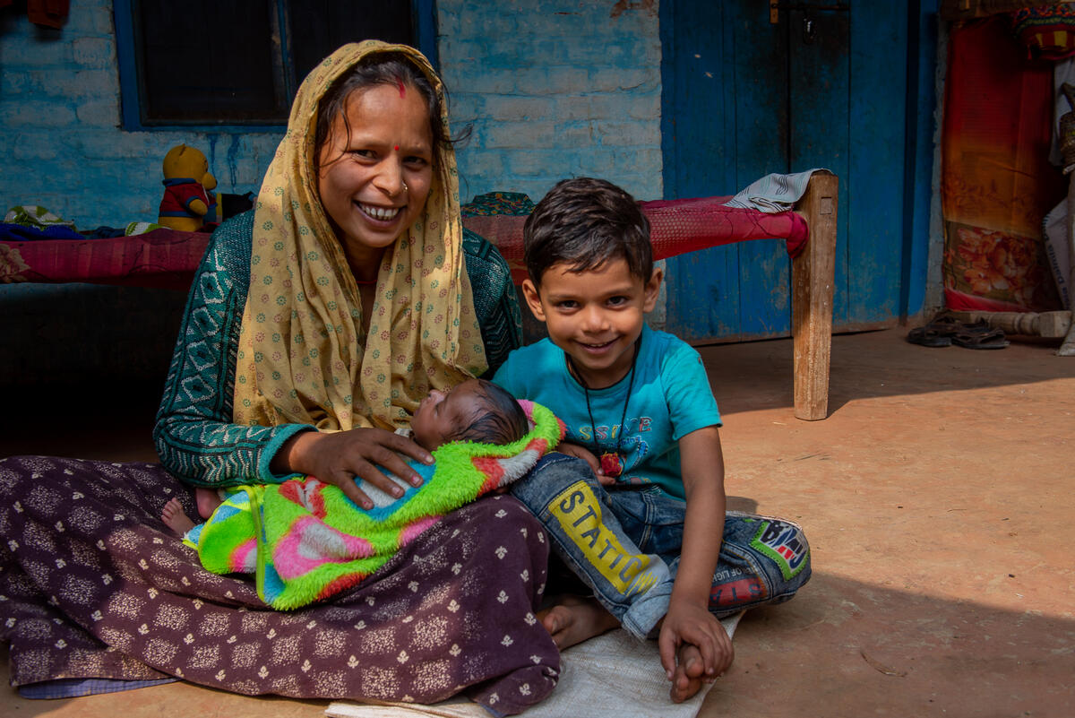 Mother sits outside holding a baby with a young child next to her