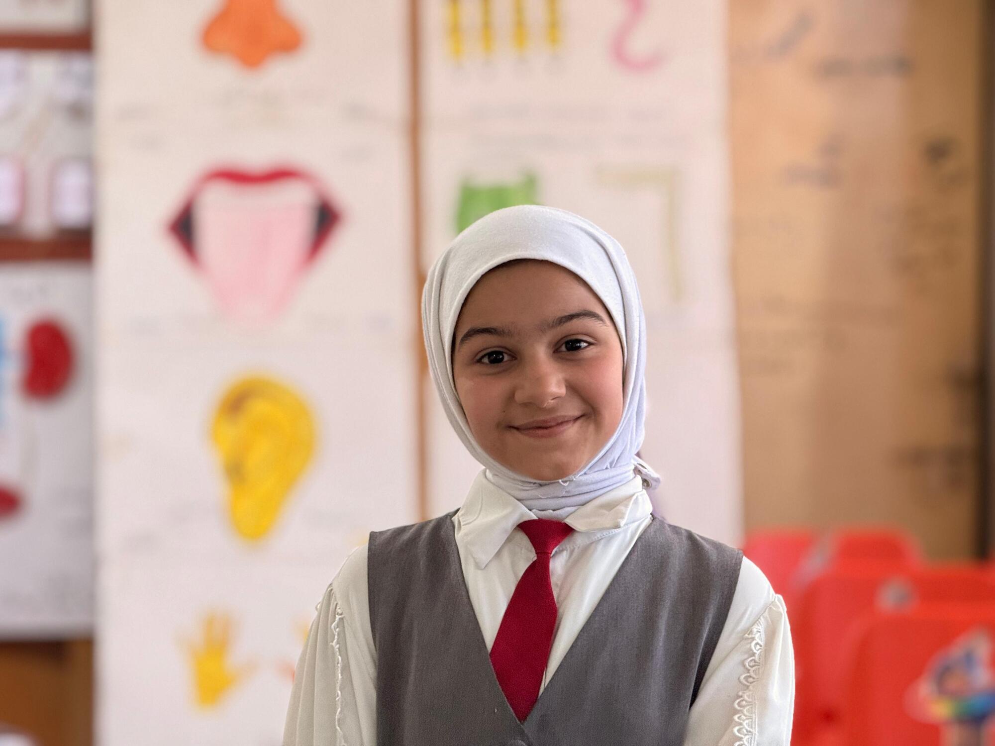 A young girl smiles at the camera while standing in a classroom.