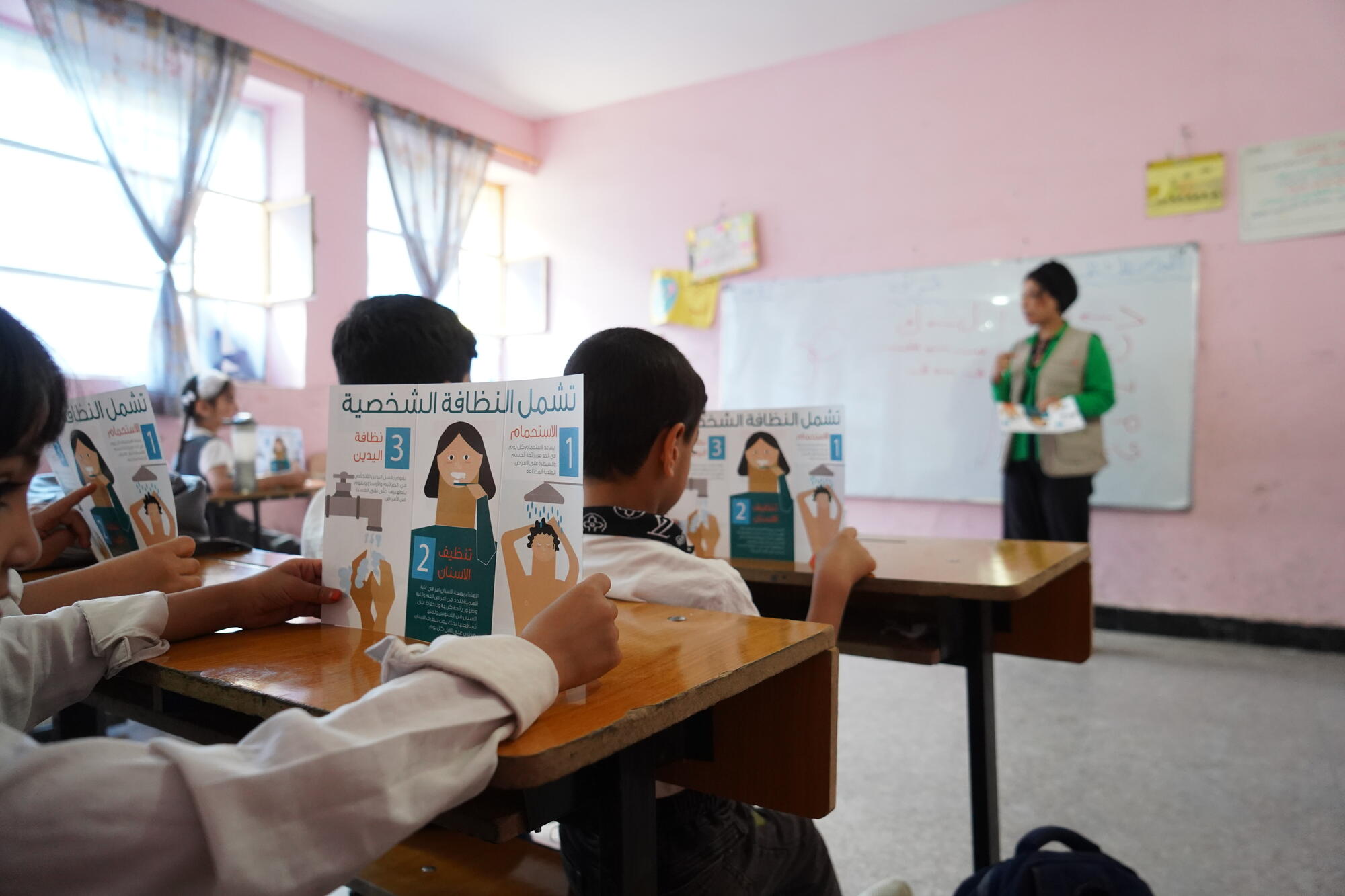 A child sitting at a desk holds up a brochure on good hygiene practices while an adult stands in front of the classroom