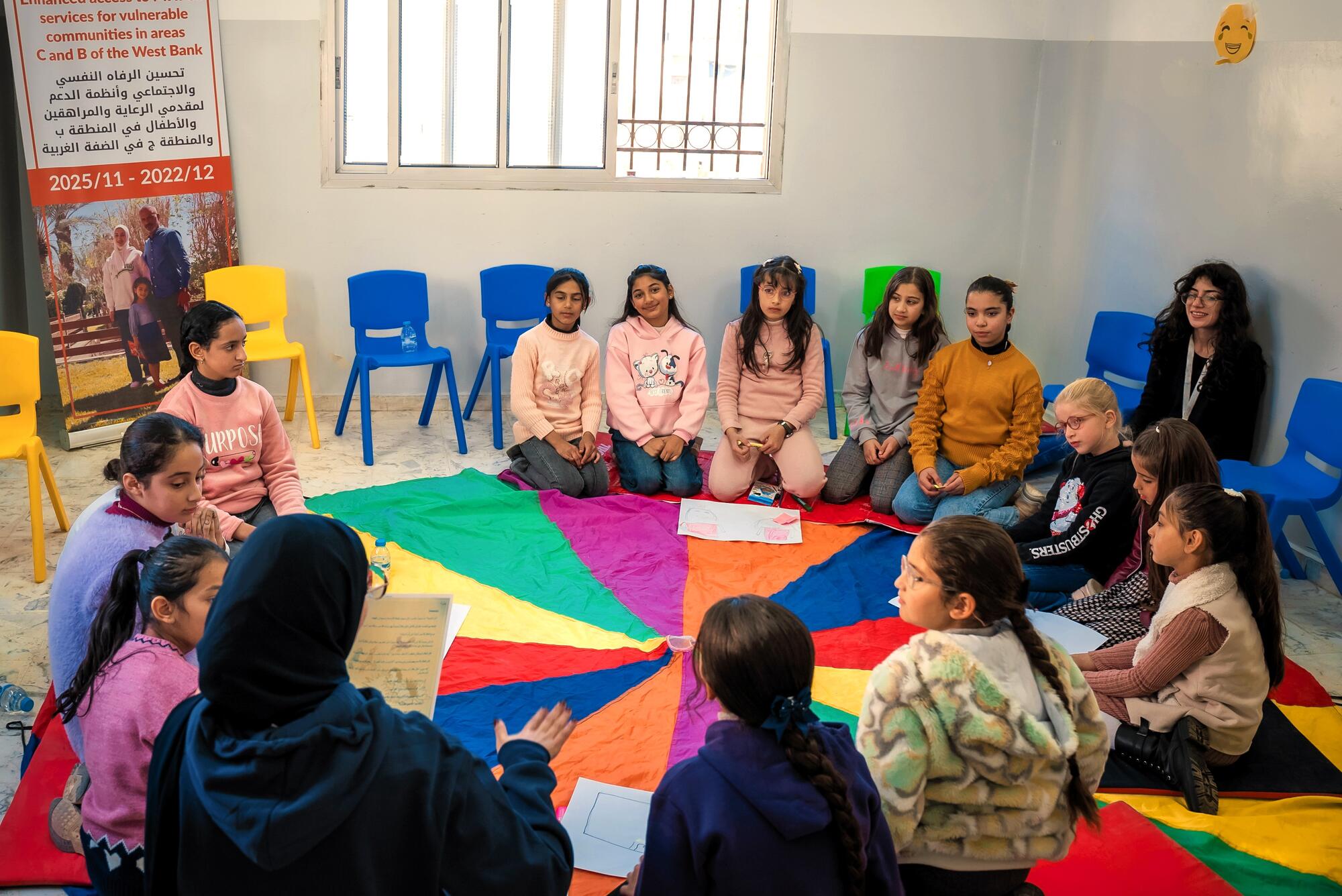 Girls sit in a circle on a bright rug in a classroom with a mental health facilitator