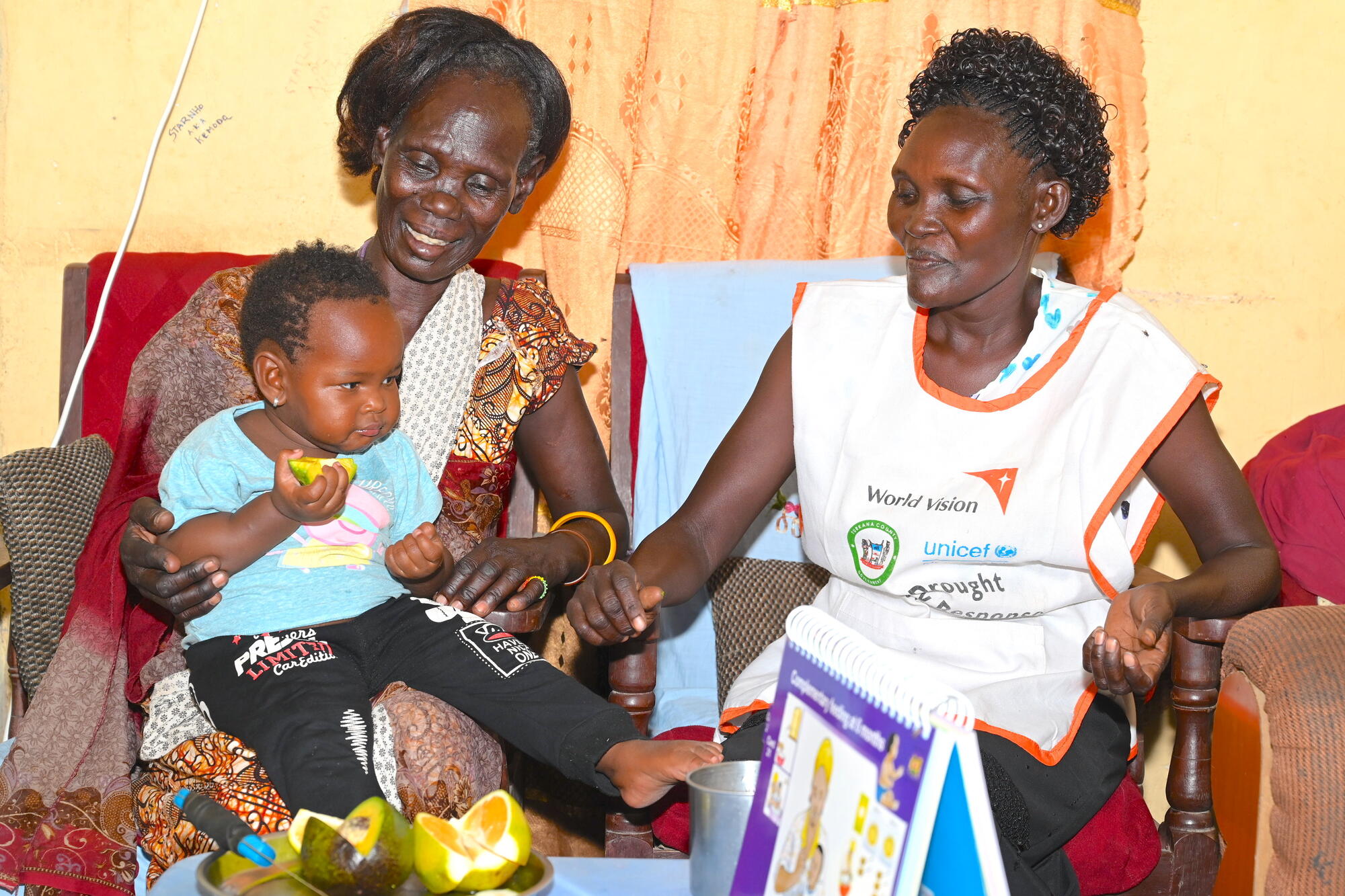 A woman wearing a World Vision vest sits with a mother and her baby who is eating a piece of food.