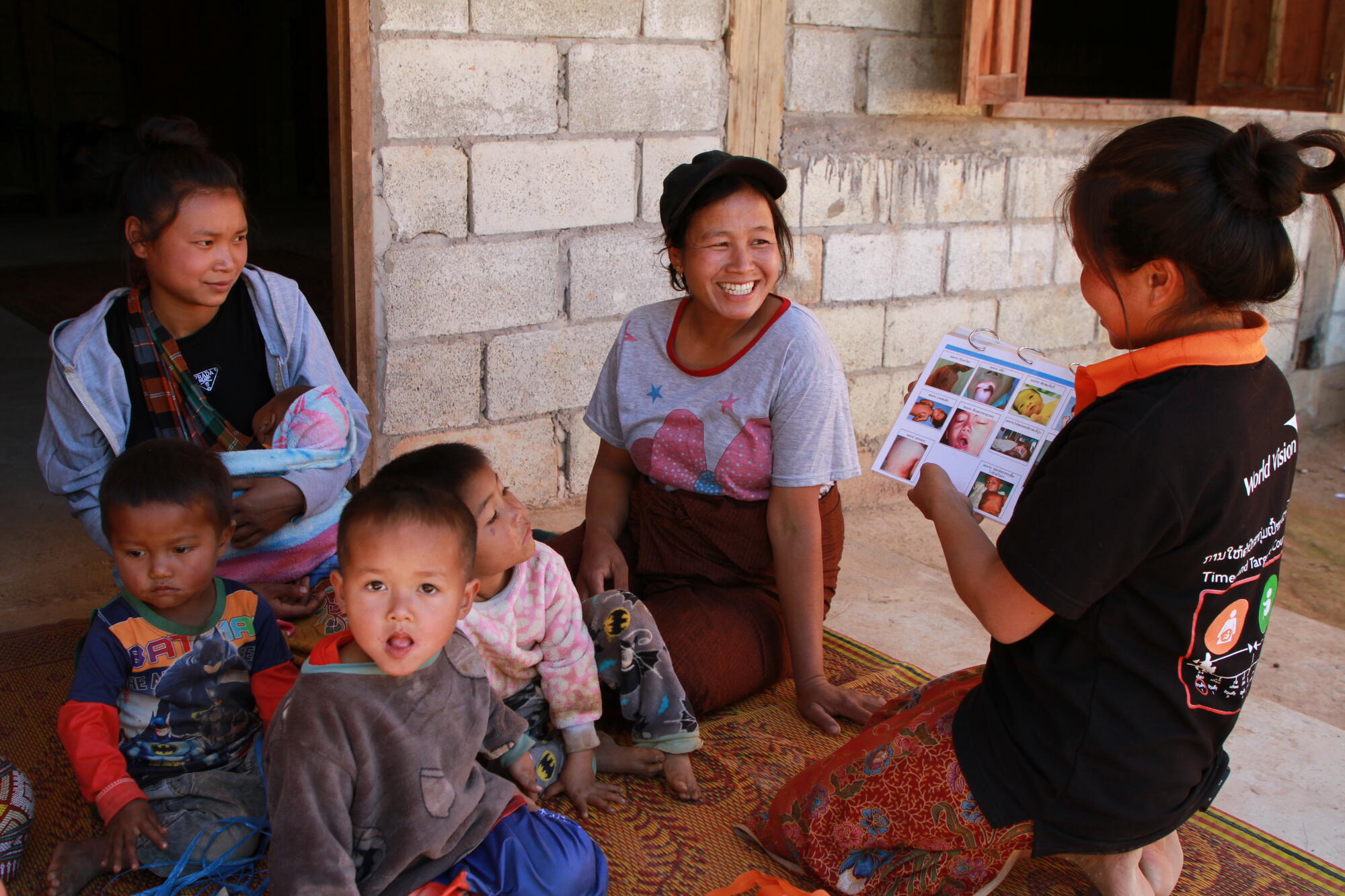 A woman sits on the floor showing a binder of health care pictures used to two mothers and their children
