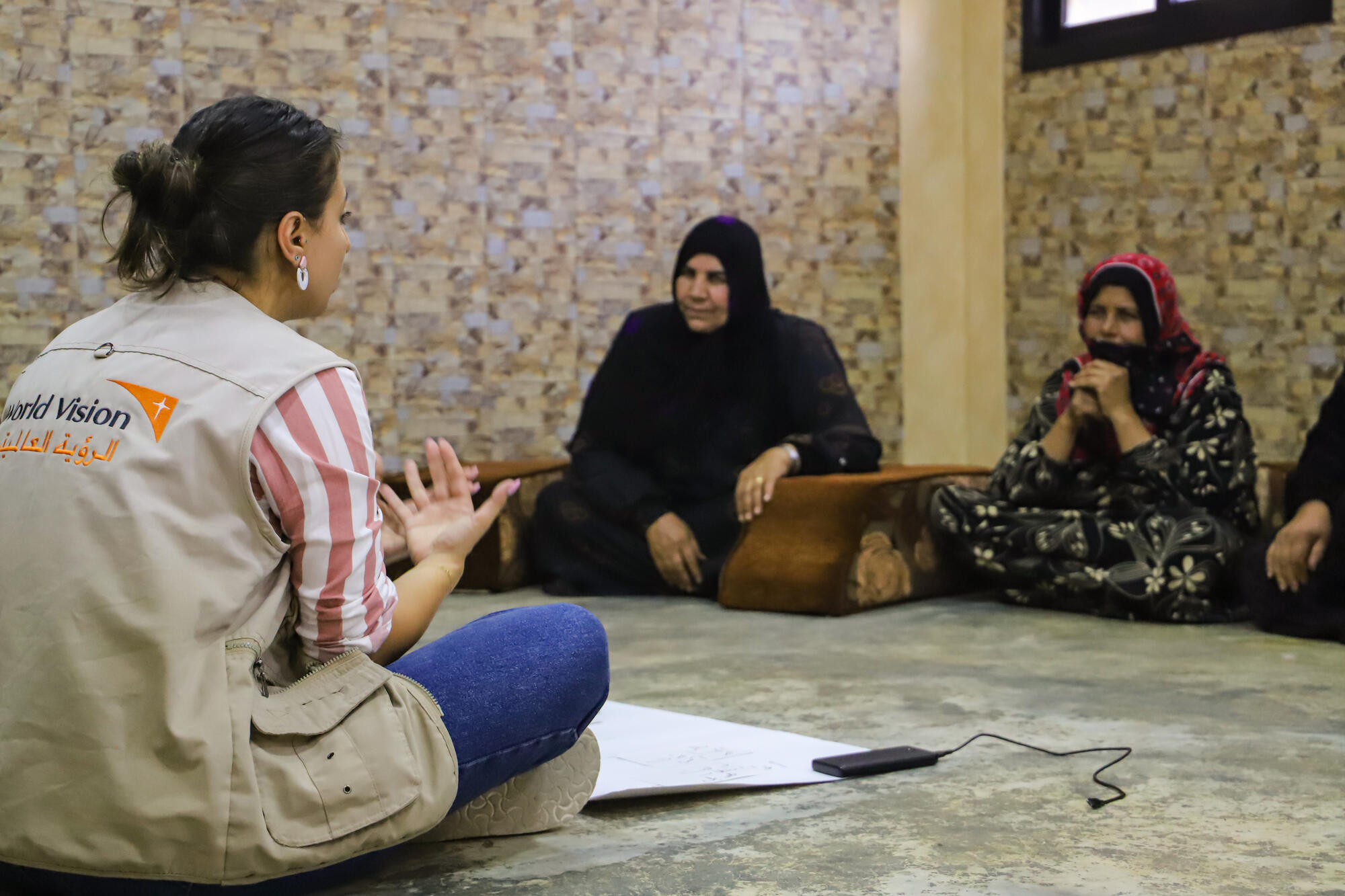 A woman wearing a World Vision vest sits on the floor speaking to Lebanese women.