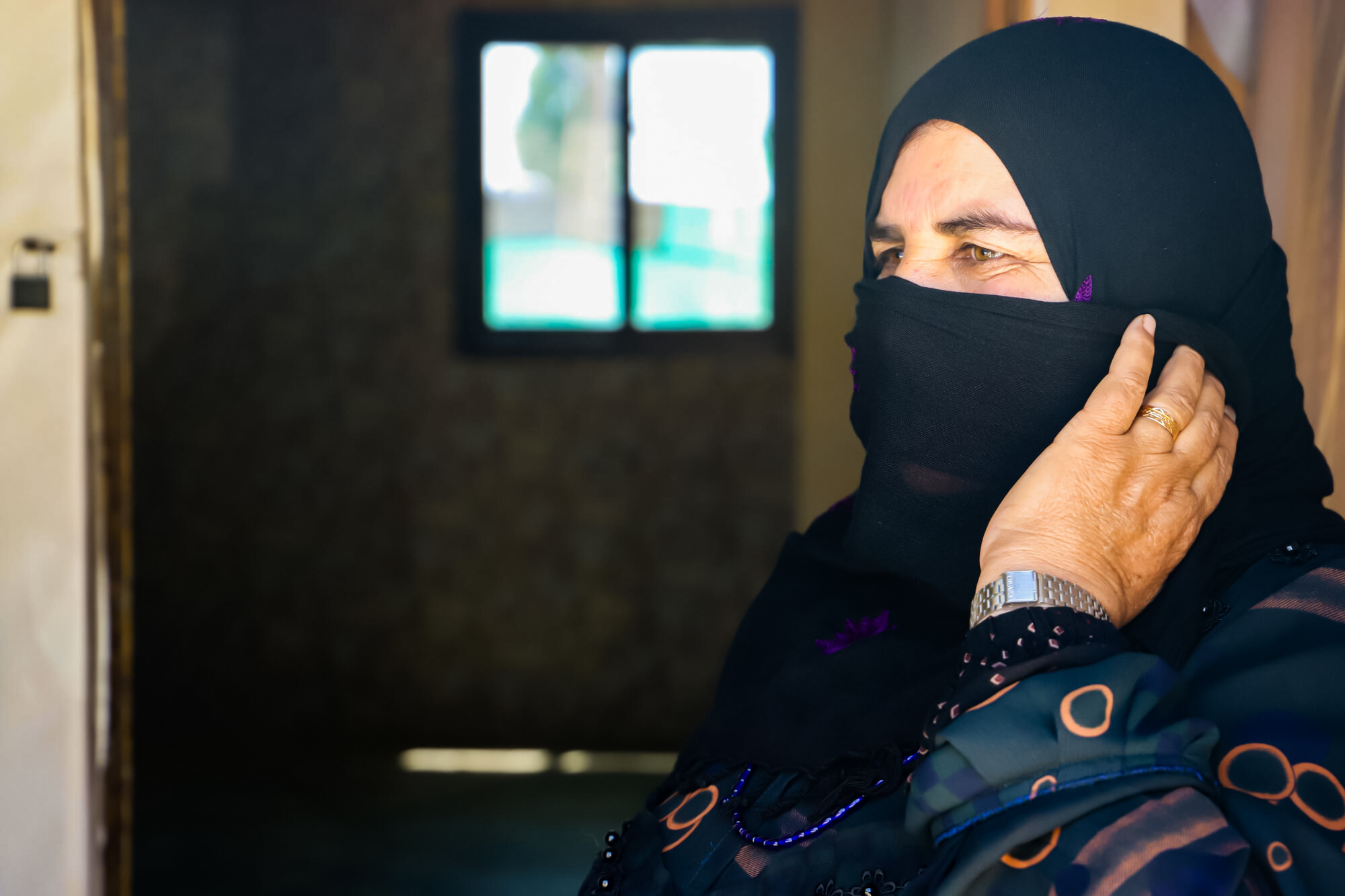 A woman wearing a black headscarf looks into the distance while sitting in front of her home.