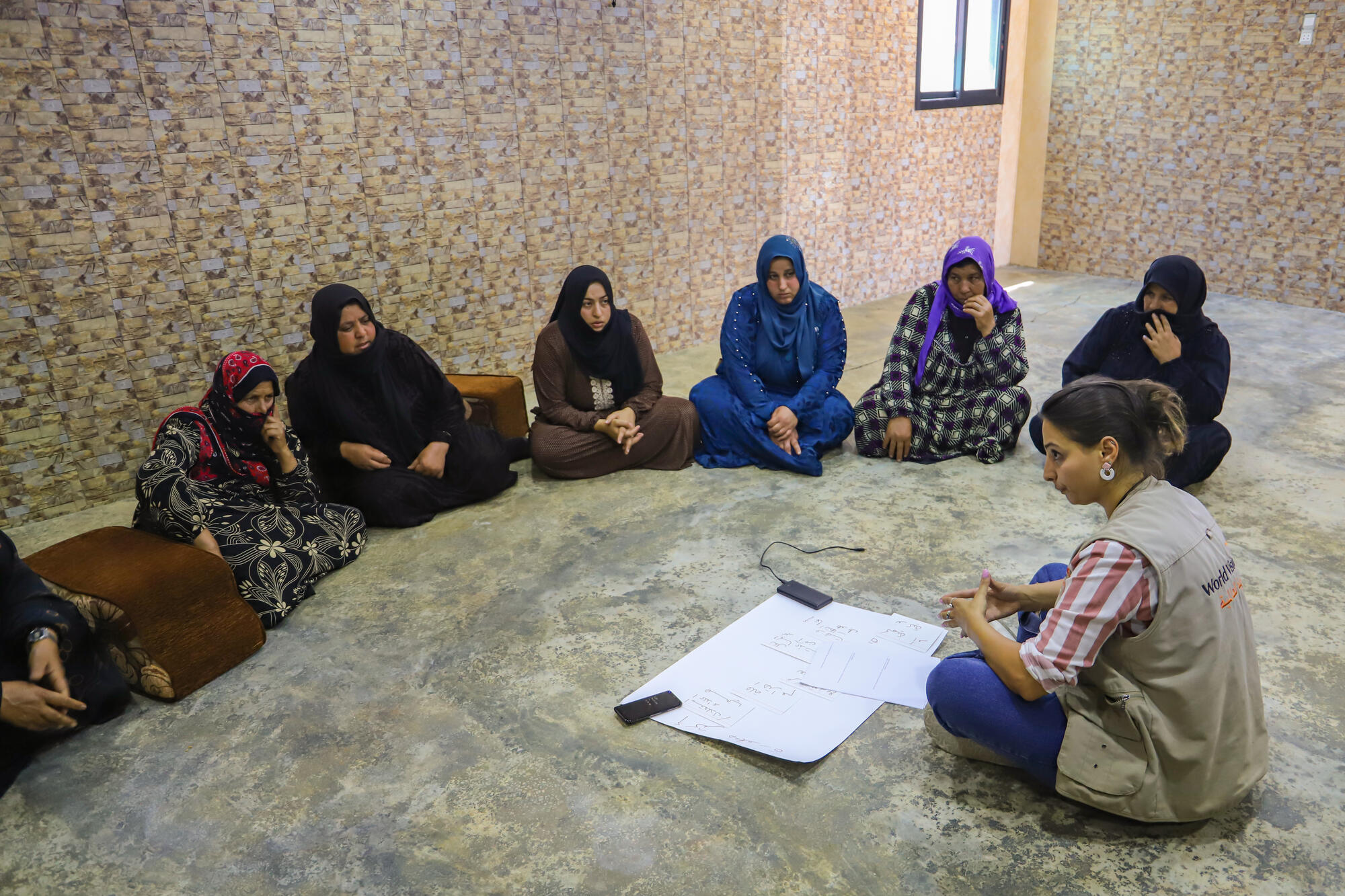 A woman wearing a World Vision vest sits on the floor speaking to 7 Lebanese women sitting in a semi circle.