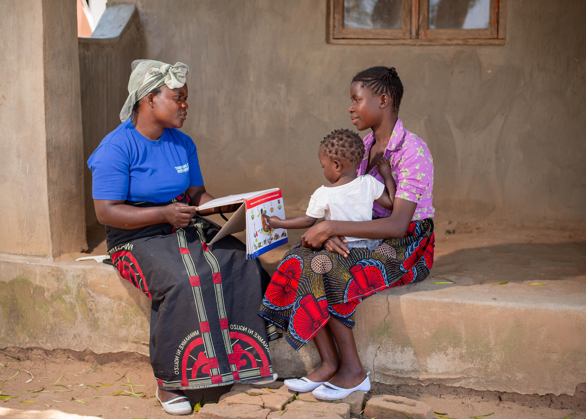 A woman hides a spiral notebook which she shows to a mother holding her child on her lap.