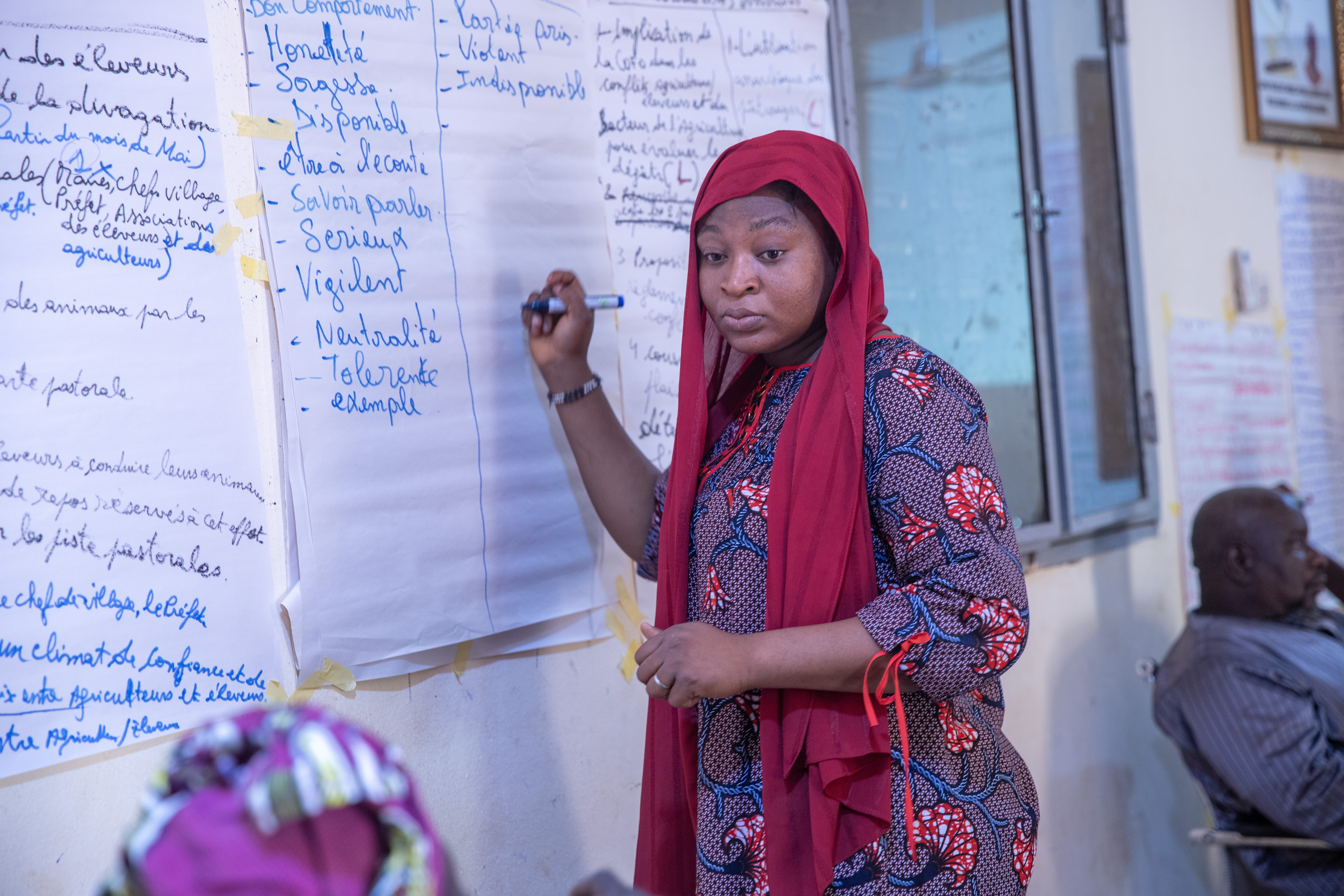 A female faith leader wearing a maroon headscarf, stands at a paper taped to a wall ready to write.