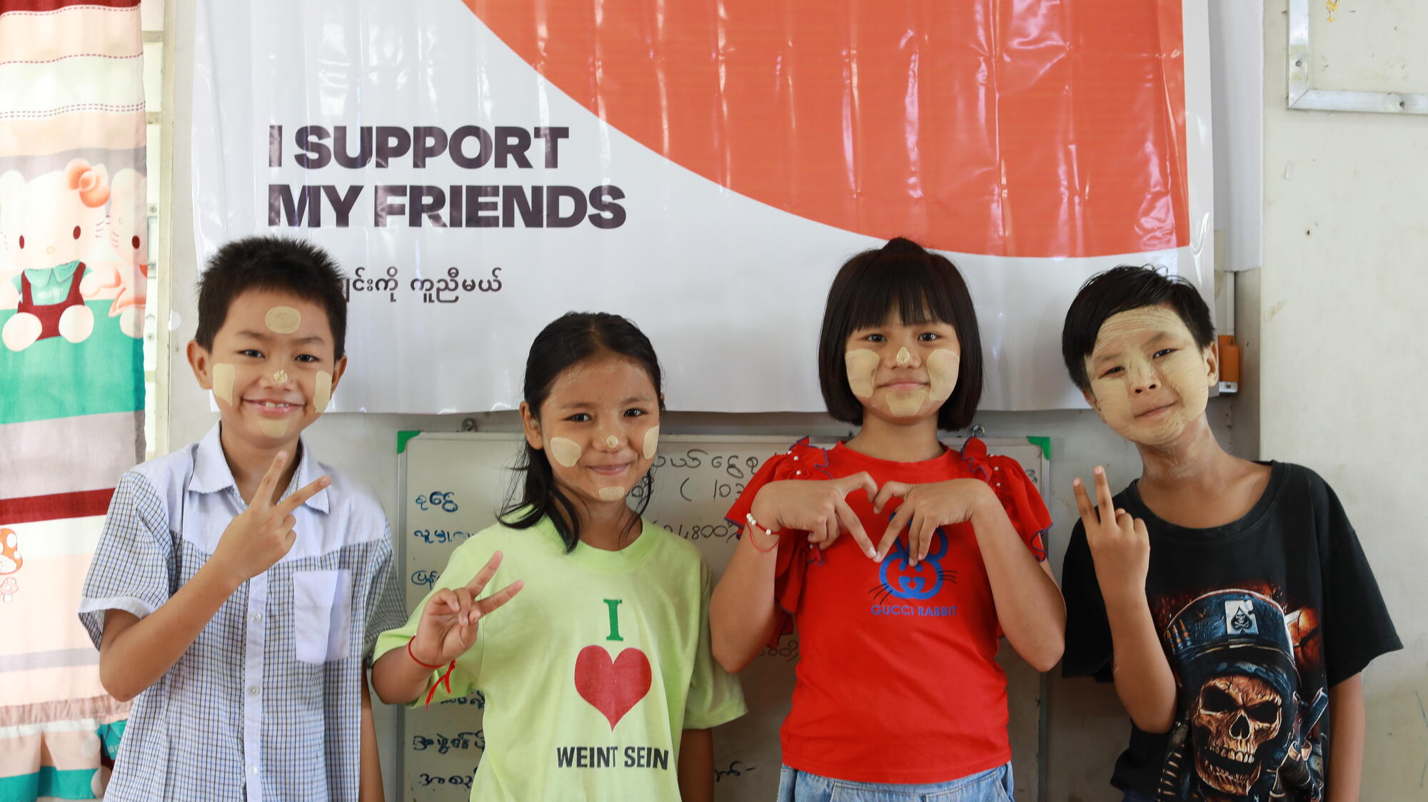 Two girls and two boys in Myanmar smile while making peace and heart signs with their hands.