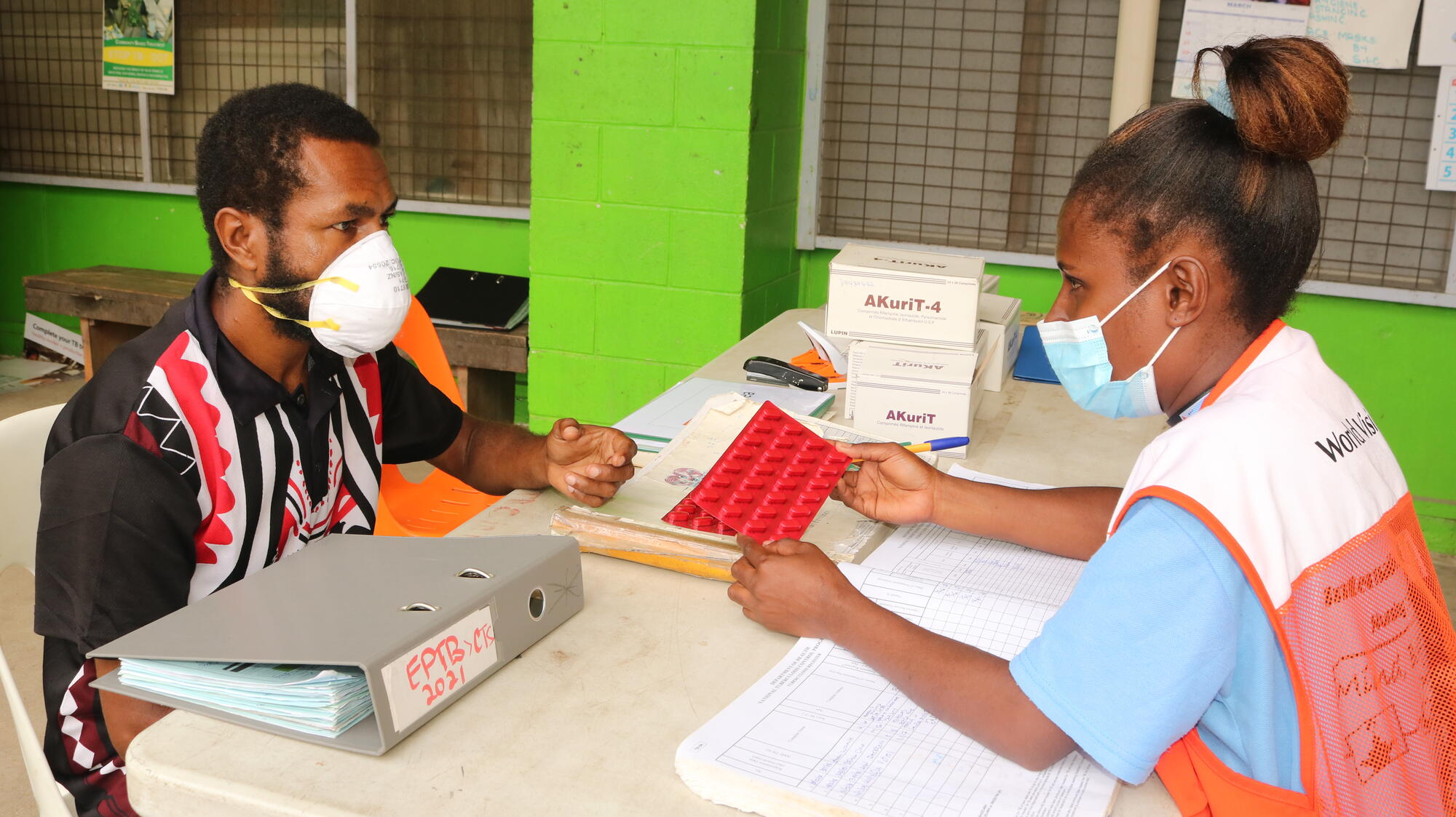 A female World Vision employee sits across the table from a TB patient.