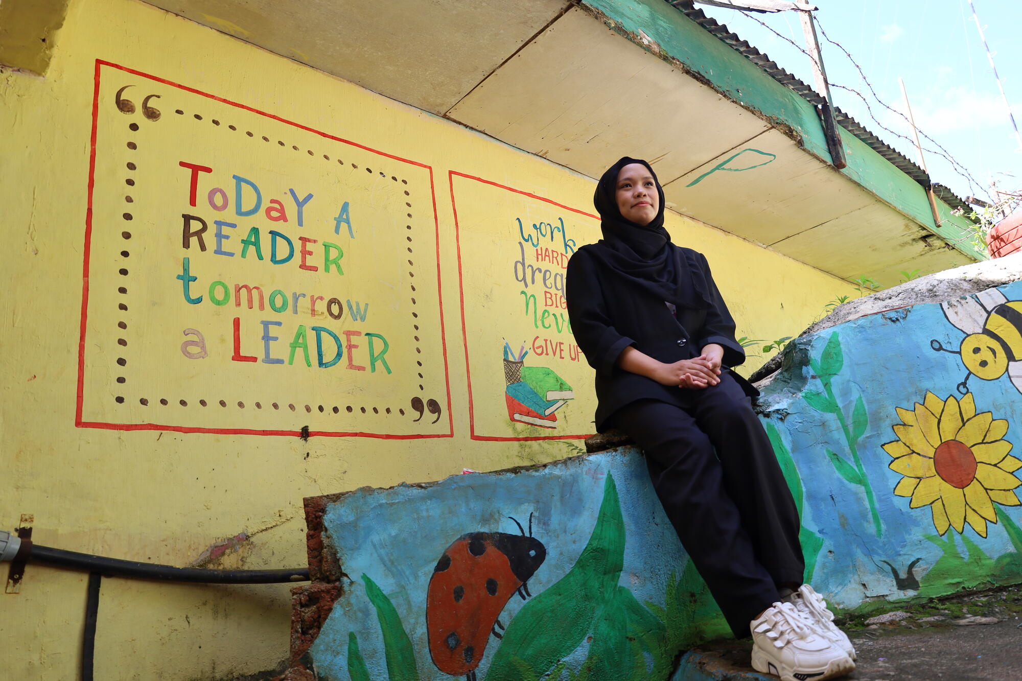 A girl sits on a low wall looking off into the distance. Behind her on the wall is a painting that reads "Today a reader, tomorrow a leader"