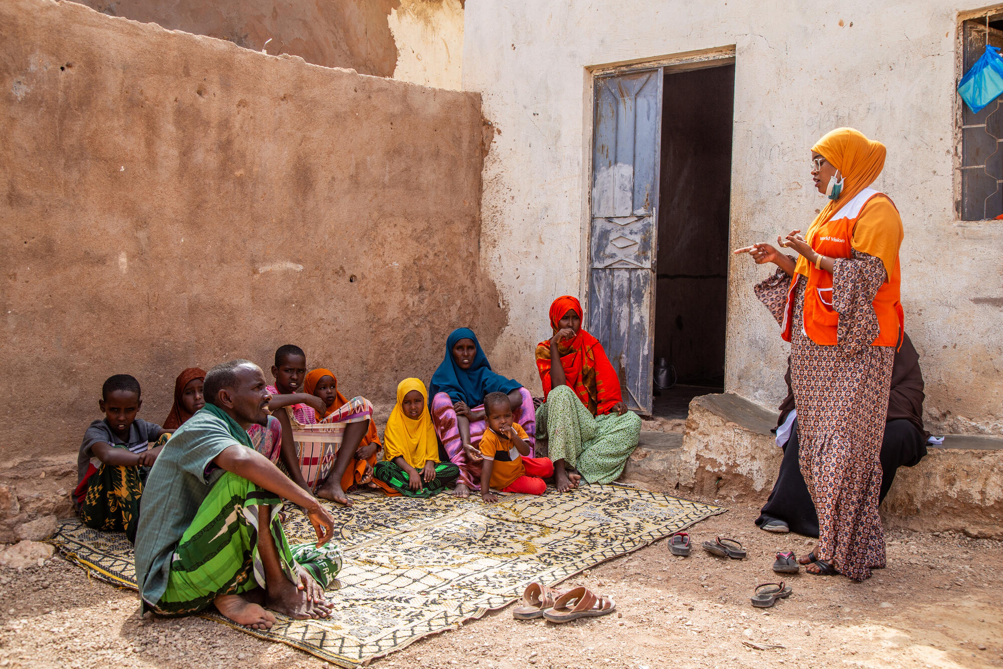 A World Vision staff member stands and talks to a family sitting on a rug outside their home.