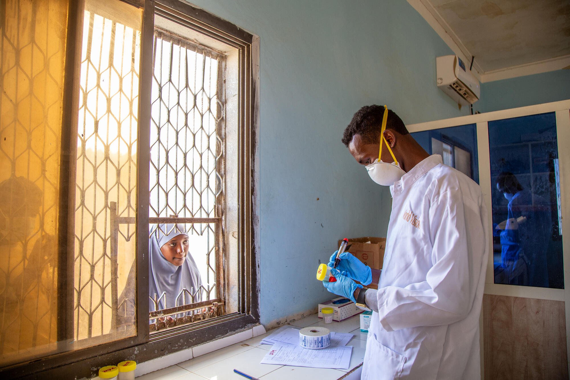 A lab technician wearing a mask writes on a sputum bin while a girl waits outside a window looking in.