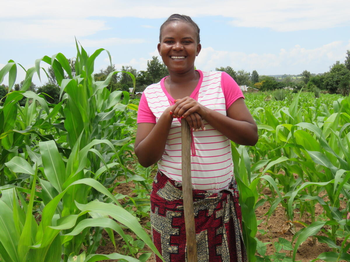 A woman in Tanzania stands in her maize harvest with a big smile.