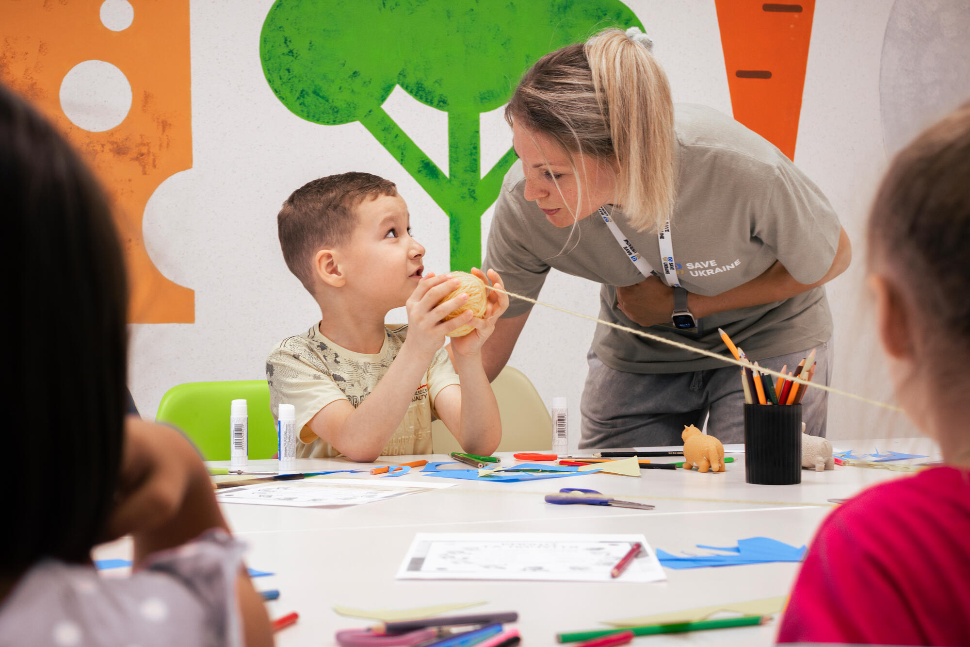 A Ukrainian Psychologist leans over a table covered in arts and craft supplies to talk to a young boy holding a ball of yarn.