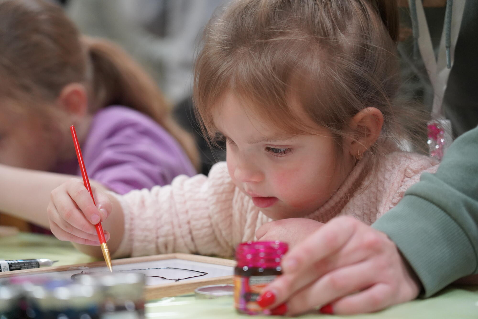 a young girl in a light pink sweater paints using a red paint brush.