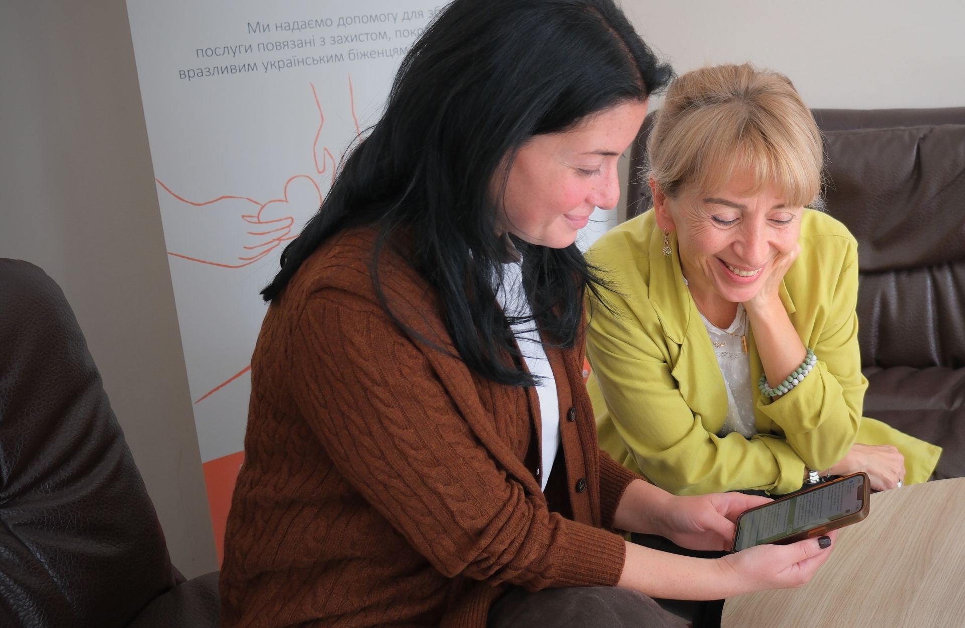 Two women sit on couches, smiling while looking at a cell phone.