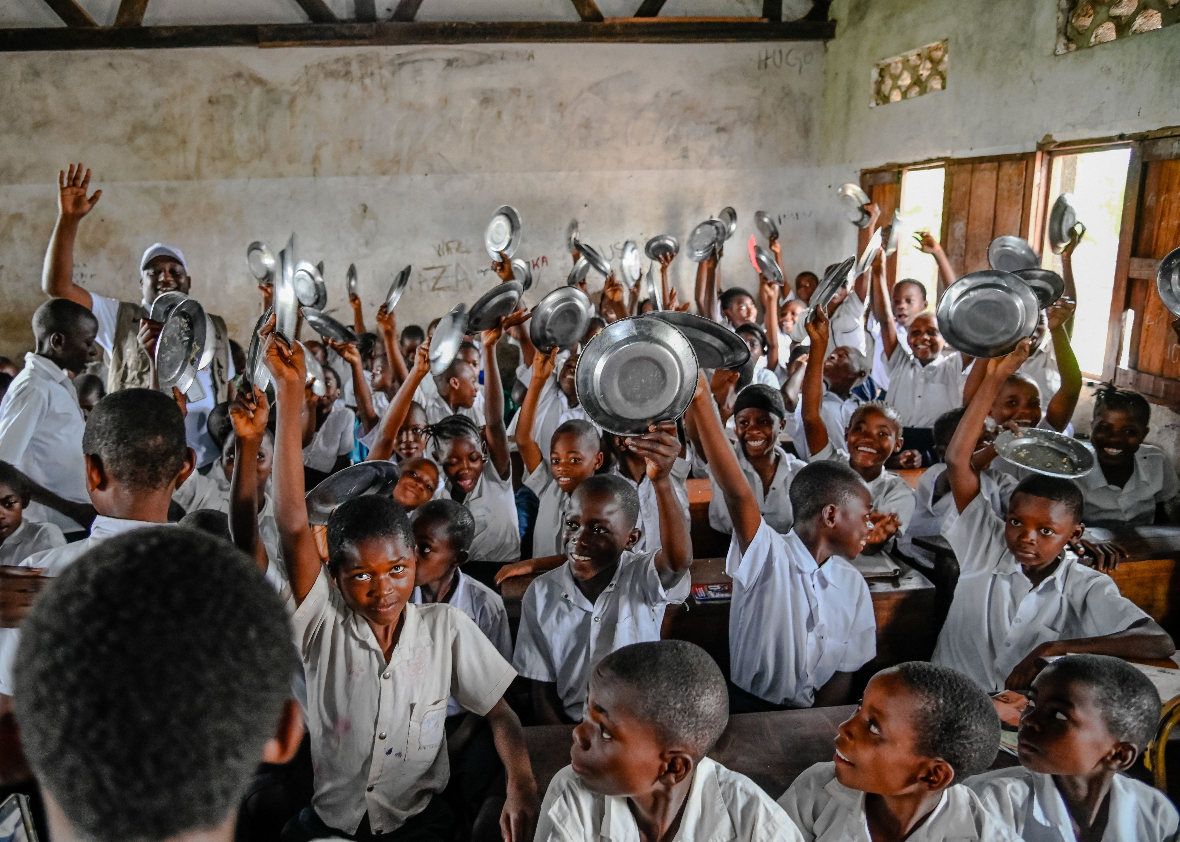 Children hold their empty bowls up in the air, ready to receive school meals through World Vision International