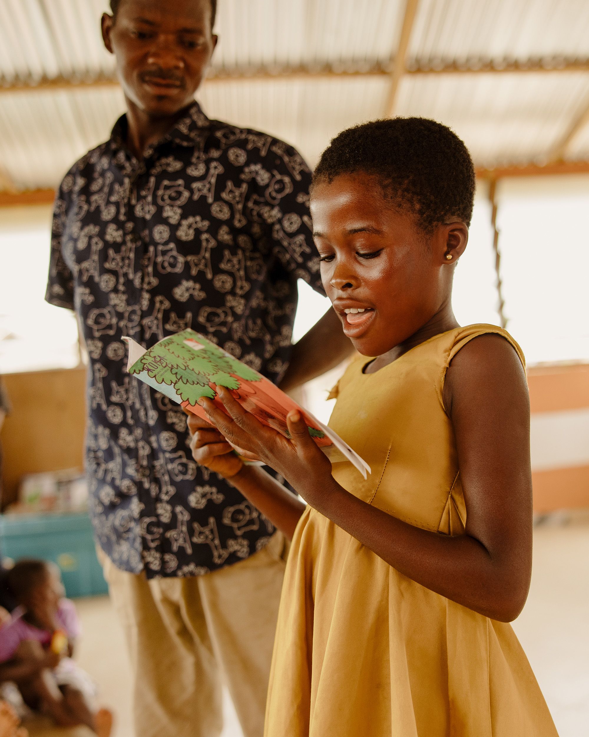 A girl in a yellow dress stands reading a book in front of a group of children