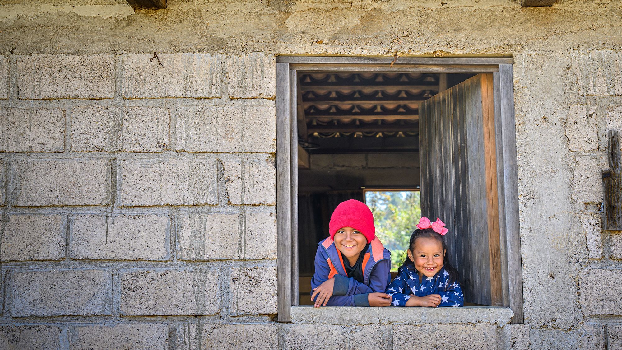 A boy and a girl smile out of a window in the middle of a grey brick wall