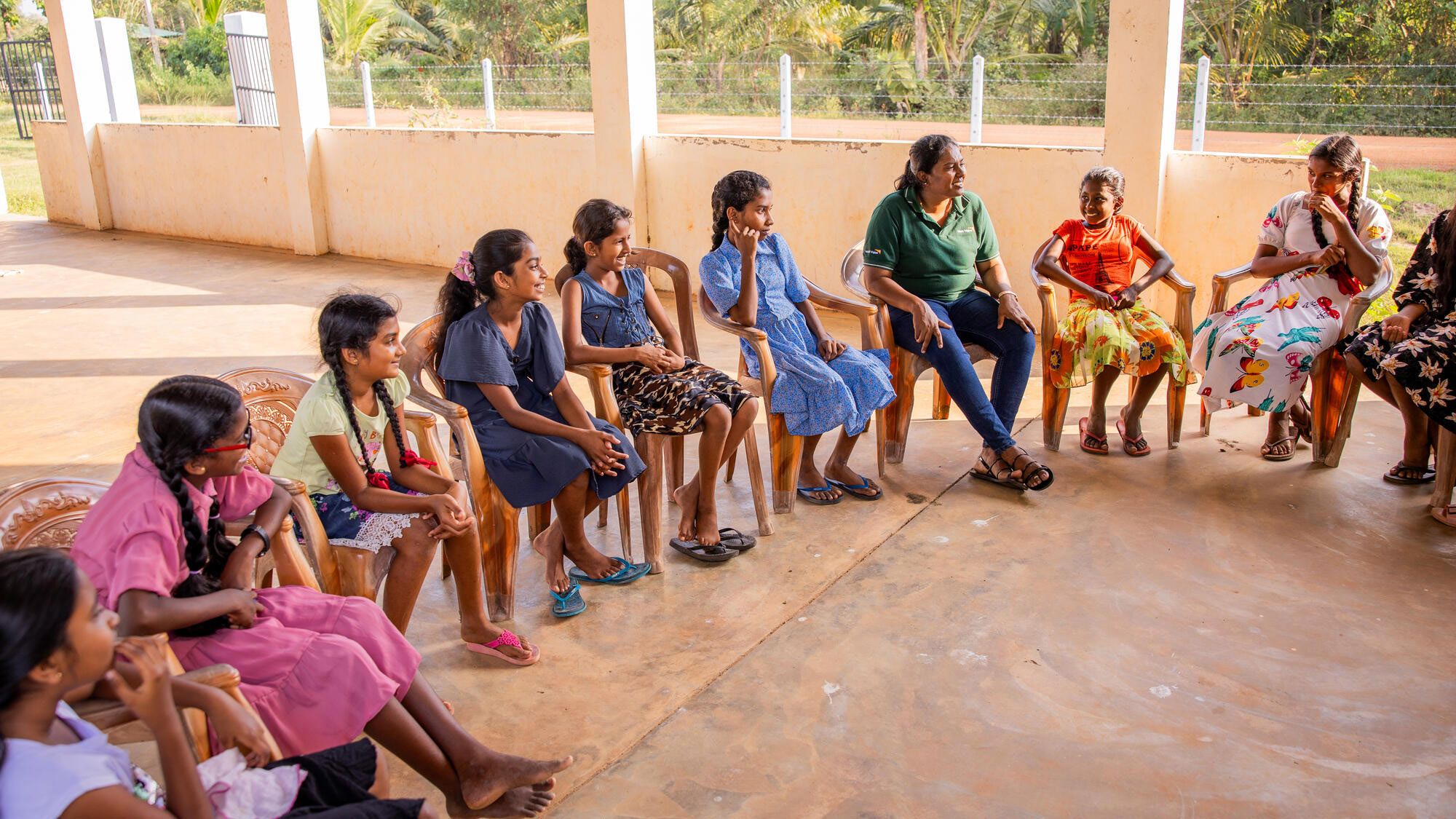 A semi circle of girls sitting in chairs with a World Vision staff member