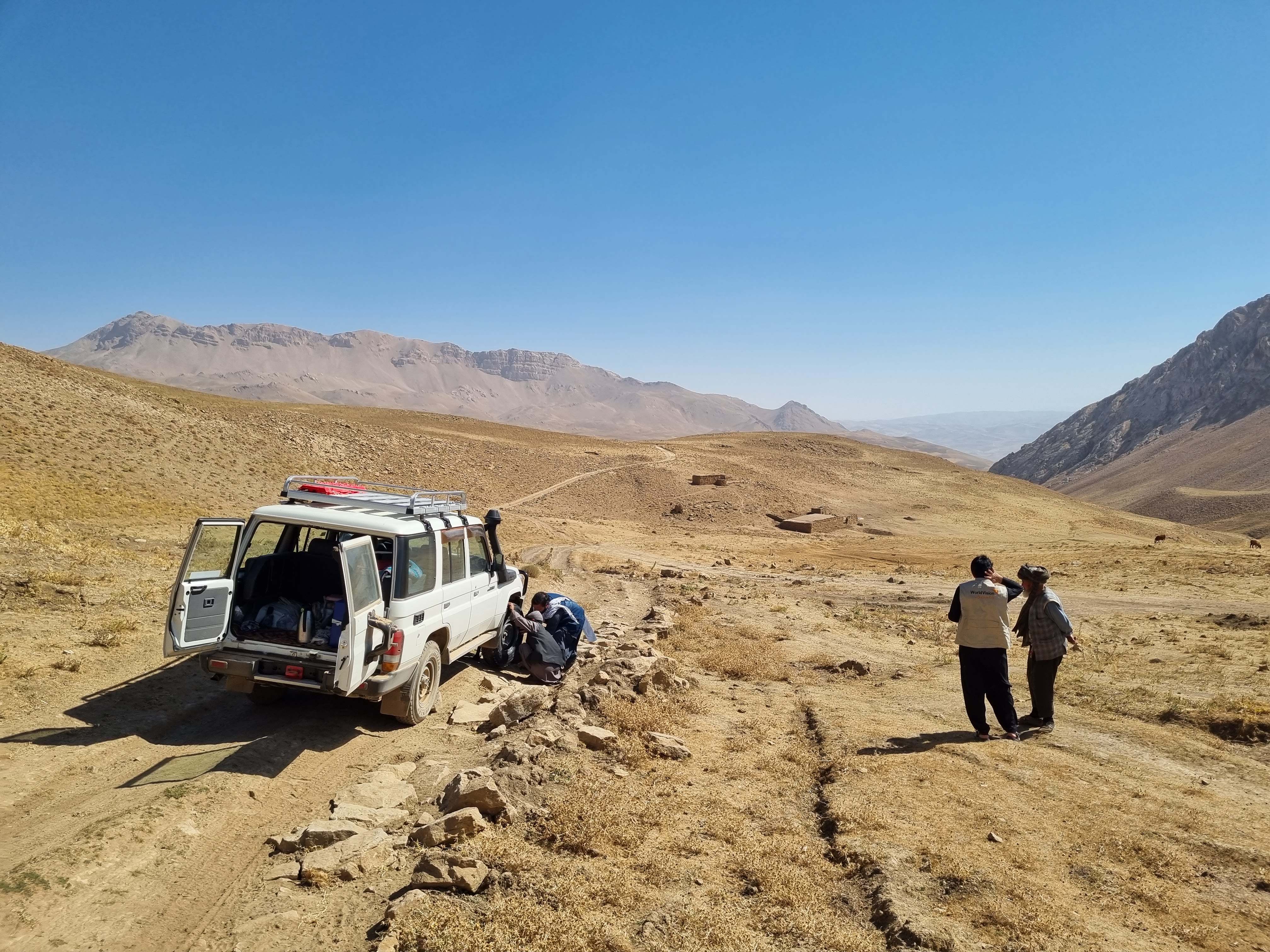 A stopped jeep having its wheel replaced while two men talk nearby.