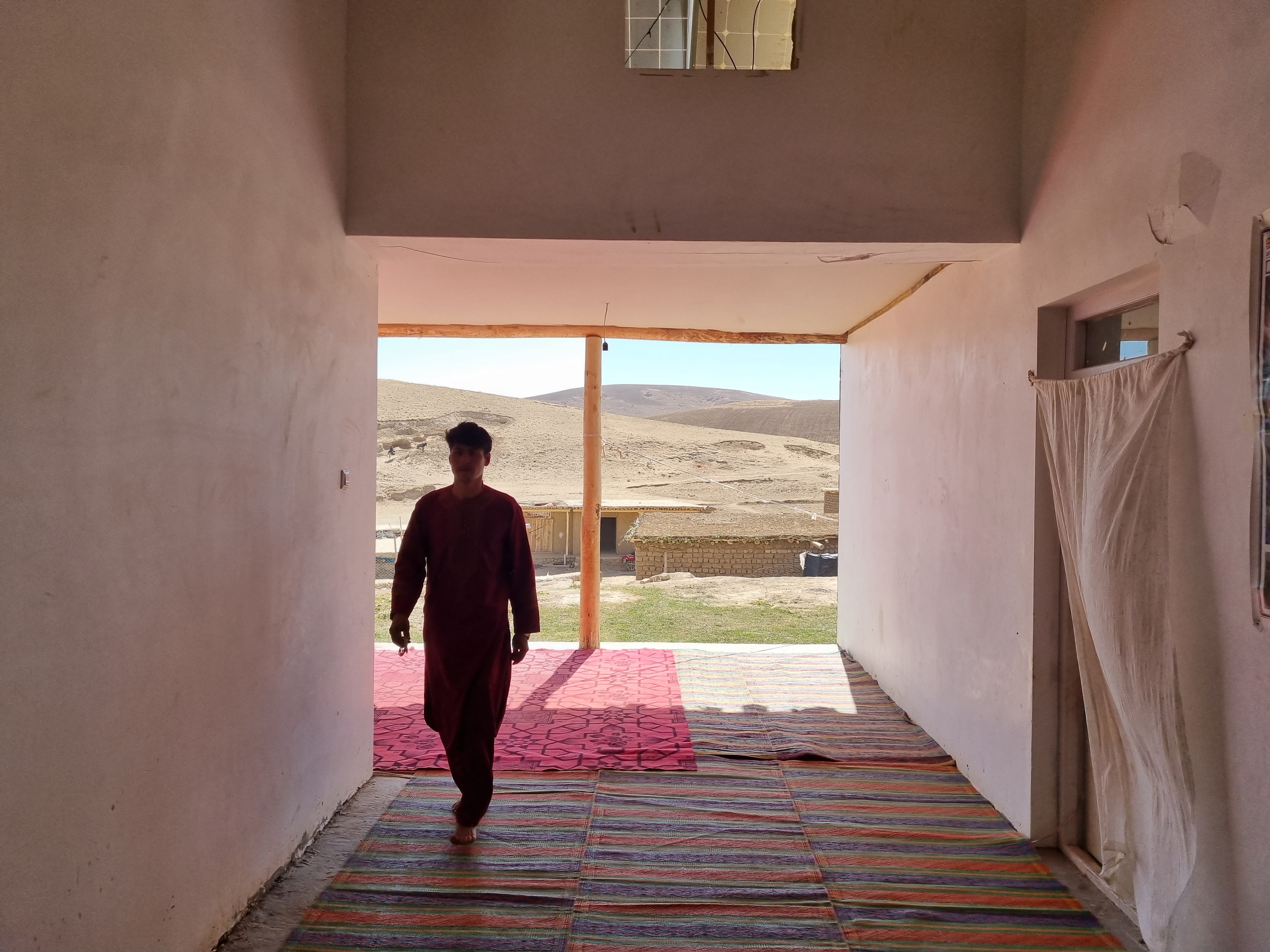 A man walking through a darkened hallway, with rugs on the floor, a sunlight exterior in the background.