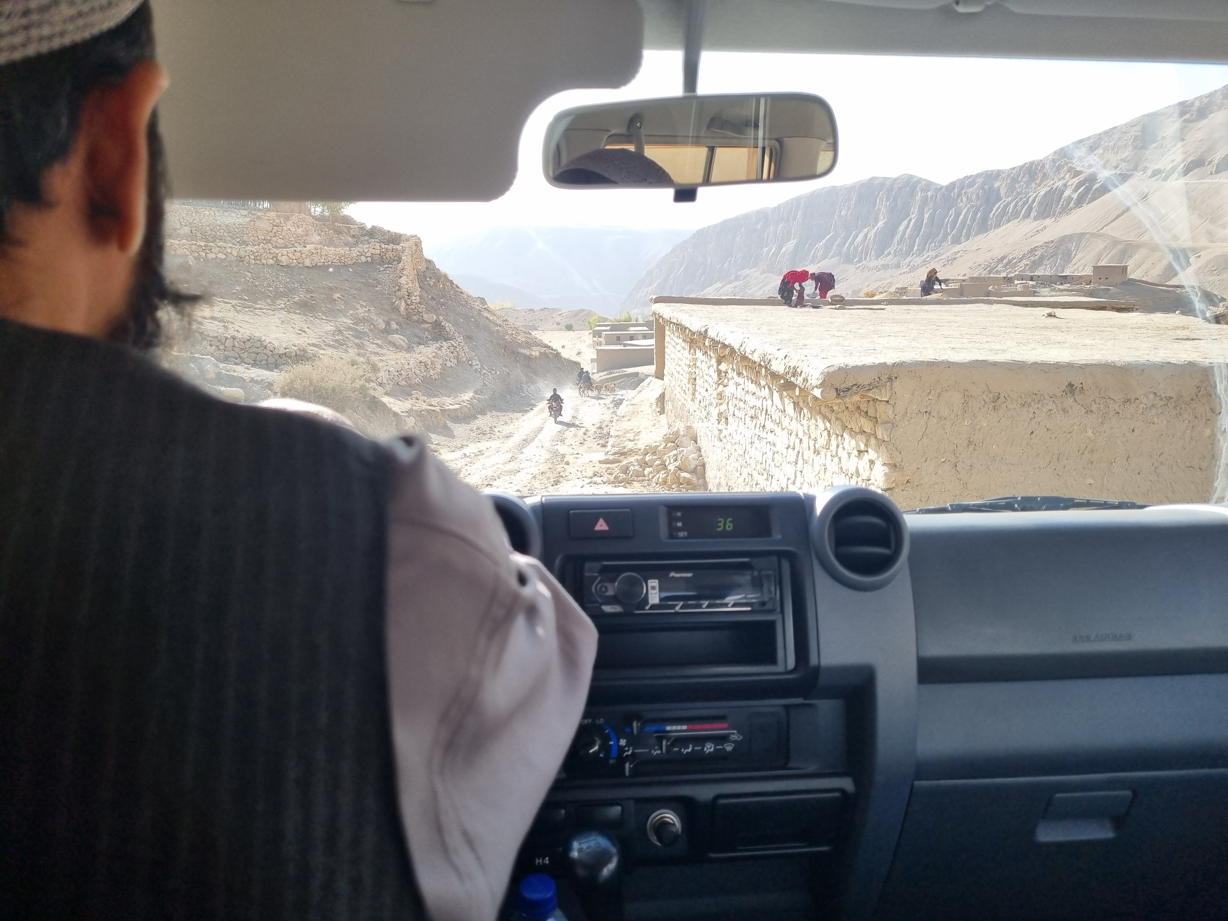 Looking through a windscreen at a dry road glimpsing women working on a roof and a man riding a motorcycle.