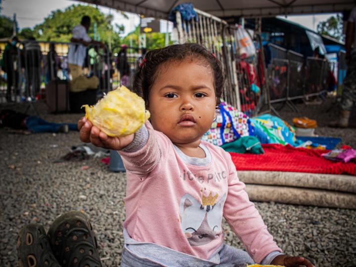 child sitting on the ground holding up a piece of food. 