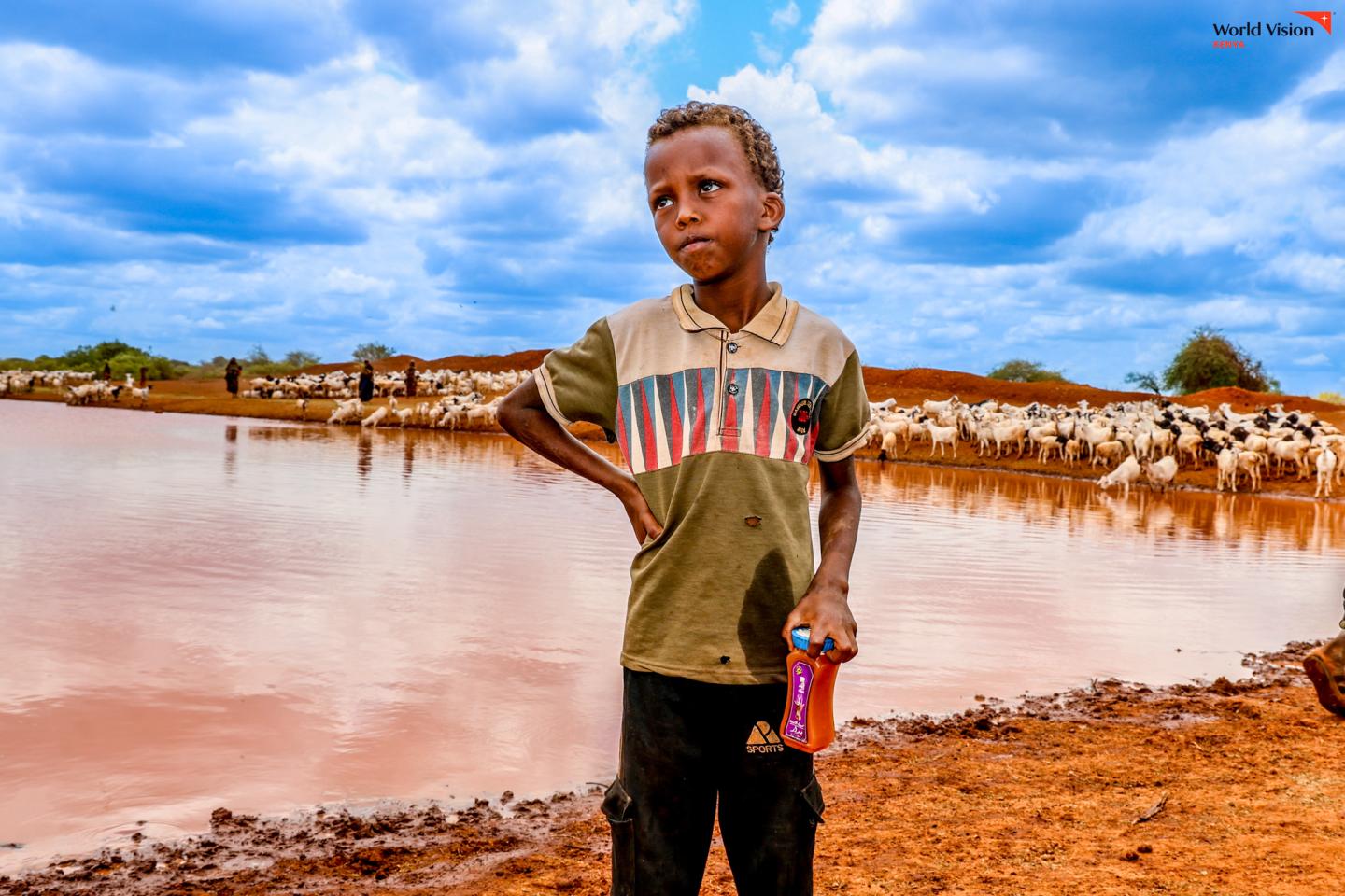 Omar, 8, stands beside a water pan in Majabow, Wajir County, holding a bottle filled with drinking water he has just collected from the water pan. ©World Vision Photo/Mirriam Kioko