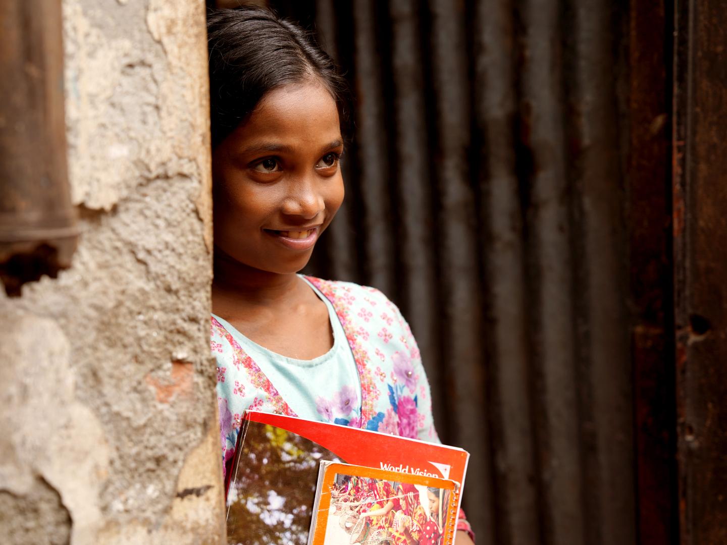 Seven-year-old Rukhsar smiles while holding her schoolbooks in the doorway of her family's home.