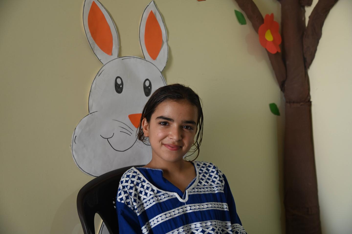 Tahani, 14, smiles while sitting inside a learning centre in Wadi Khaled, Akkar, with colourful handmade decorations behind her.